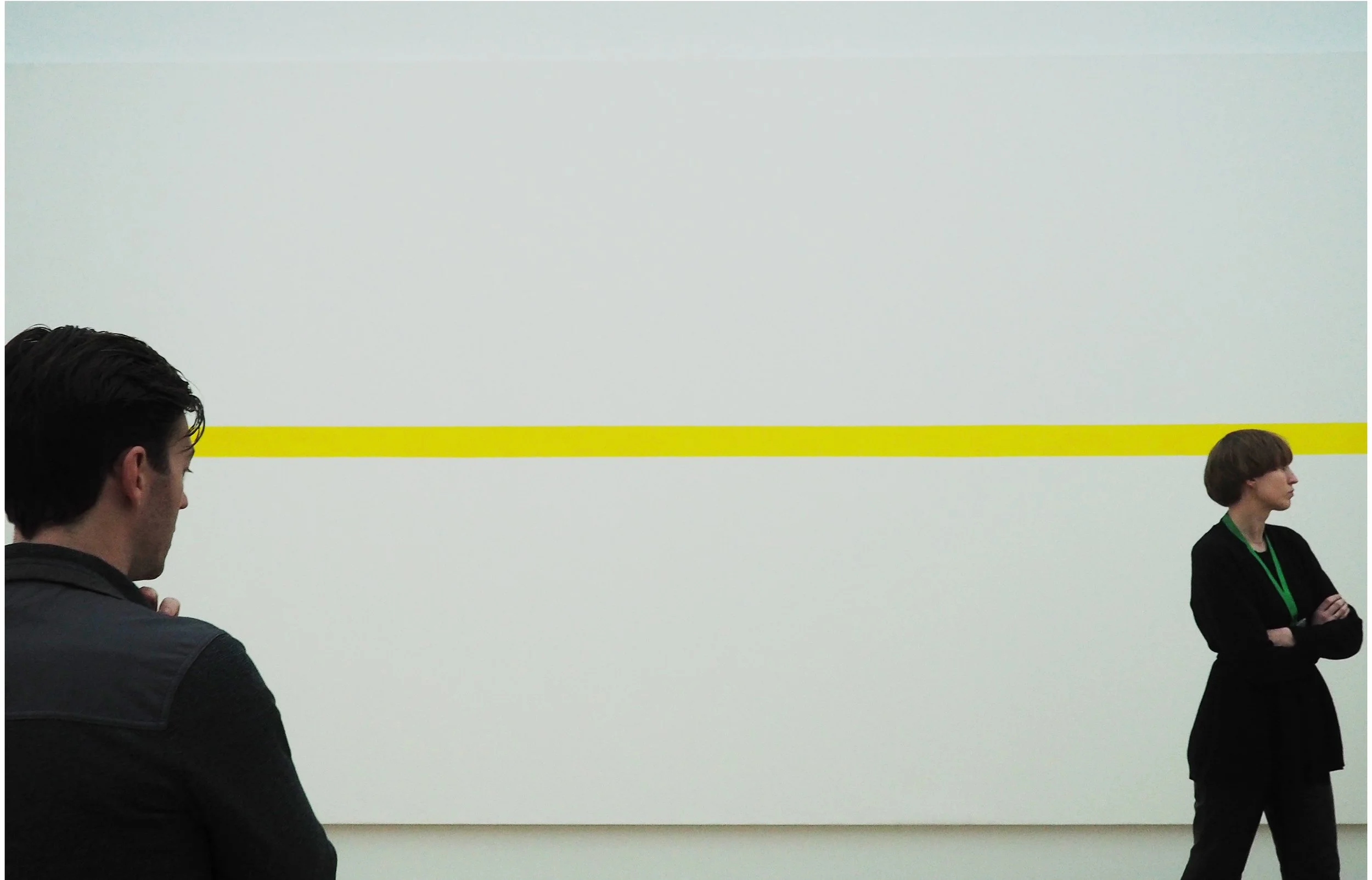 A man and a woman stand apart in front of a large white wall with a horizontal yellow line.