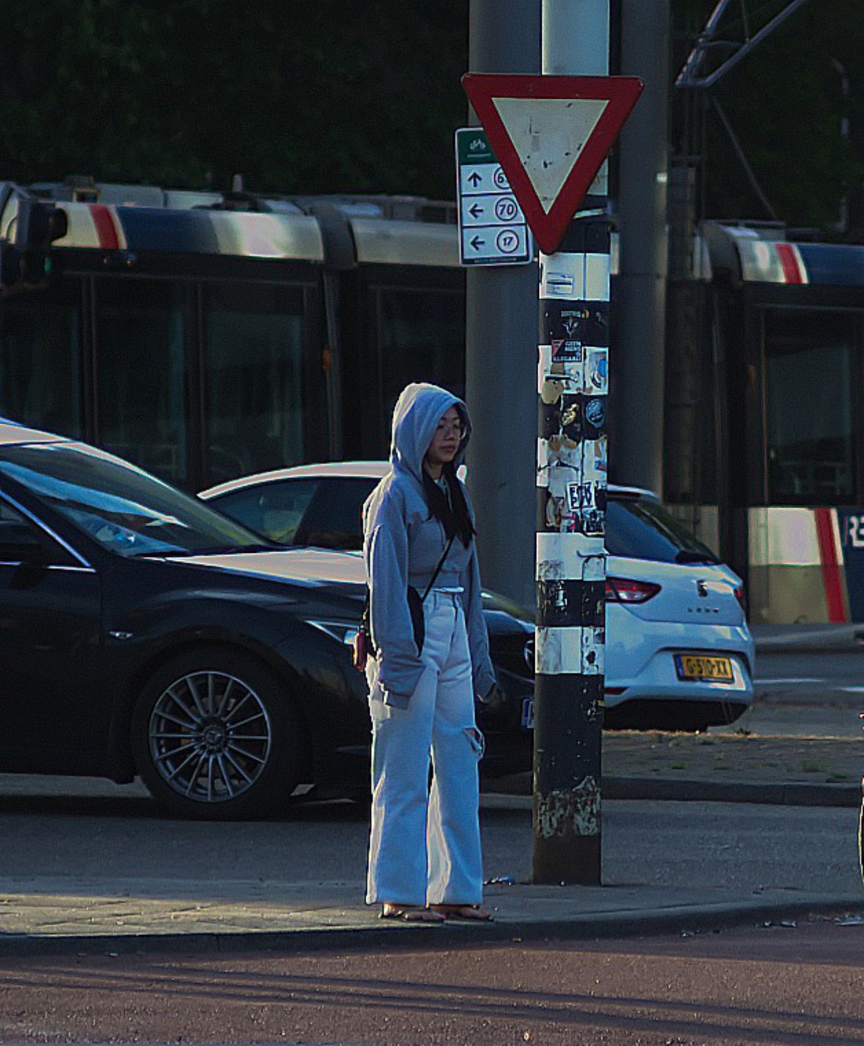 A woman in a light gray hoodie and white pants standing on a sidewalk near a pole with stickers and signs, with cars and buses in the background.