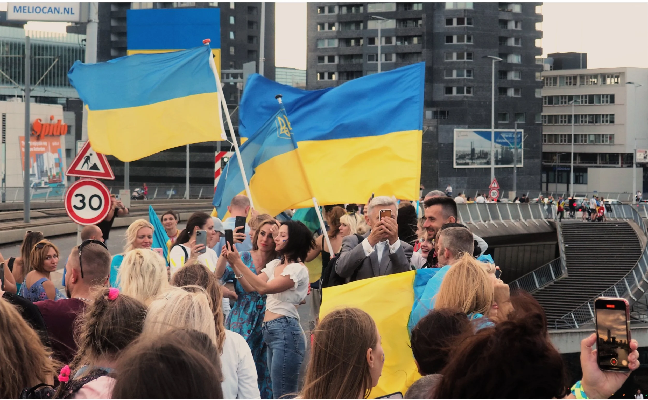 Crowd of people at a protest or gathering, holding Ukrainian flags with yellow and blue colors, in an urban area with high-rise buildings.