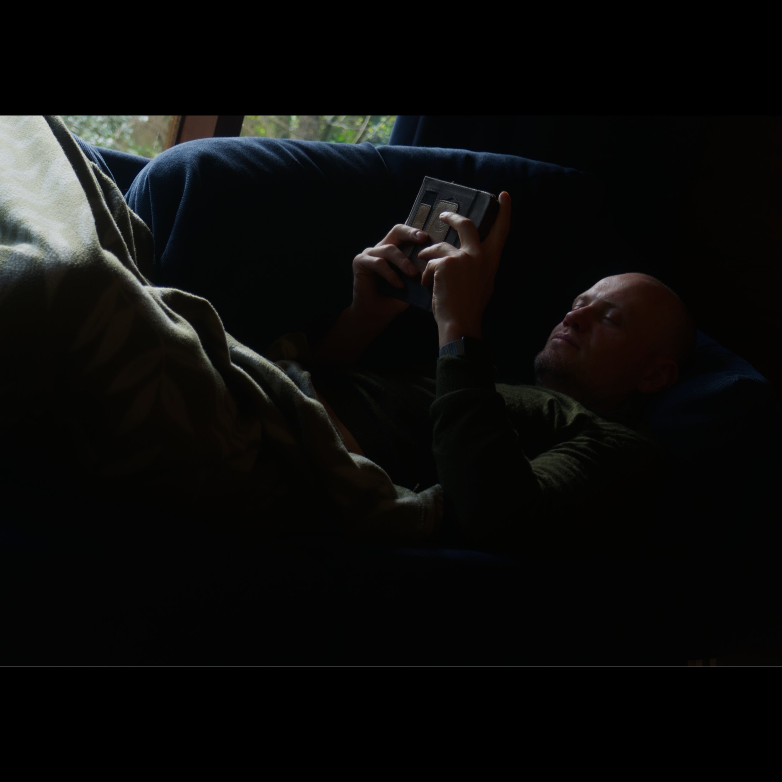 A man lying on his back on a bed, using a smartphone, with a window showing greenery in the background.