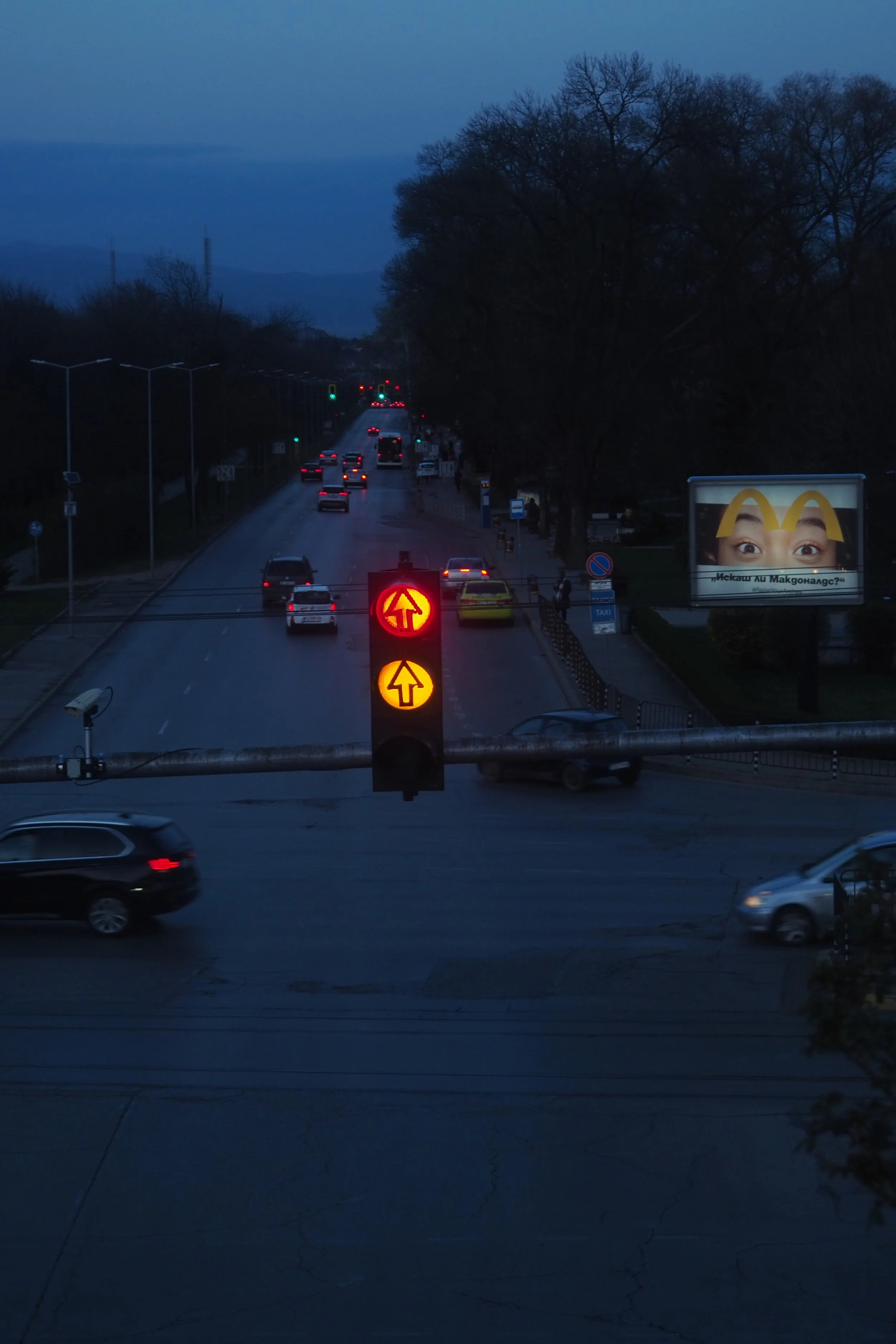 Traffic light showing red for straight and green for turning left, with cars on the road and a billboard advertising McDonald's in the background during dusk.