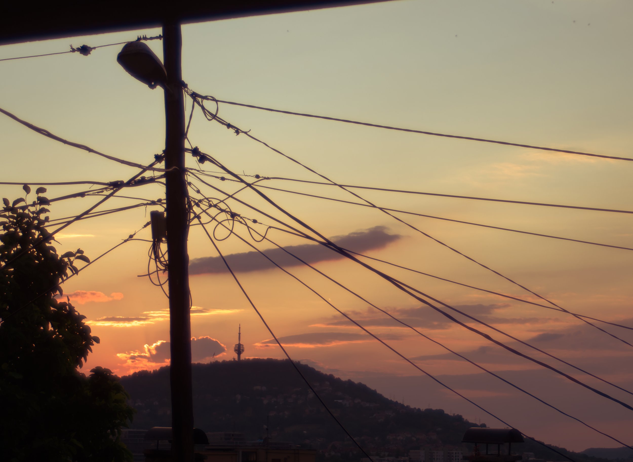 Silhouette of a utility pole with tangled wires against a sunset sky with clouds and a distant hill.