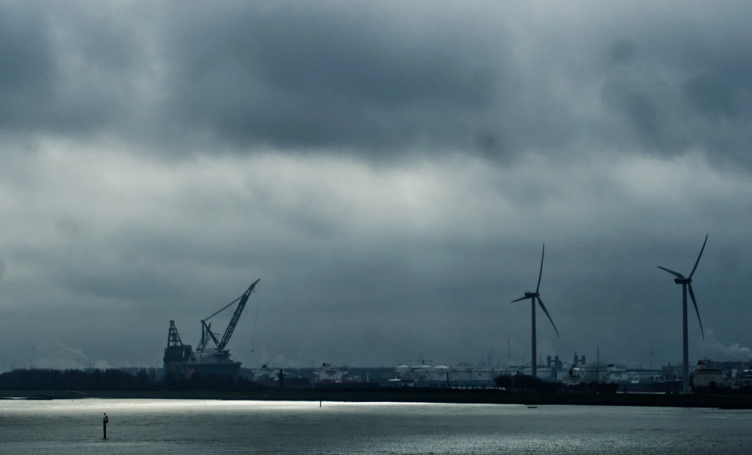 A seascape showing an industrial port with cranes, wind turbines, and ships under a cloudy sky.