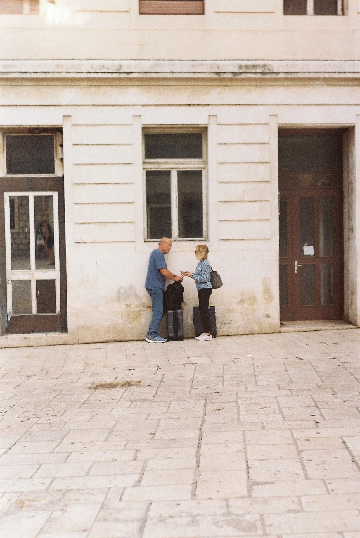 A man and woman exchanging contact details on a city sidewalk outside a building, both standing beside luggage and backpacks.