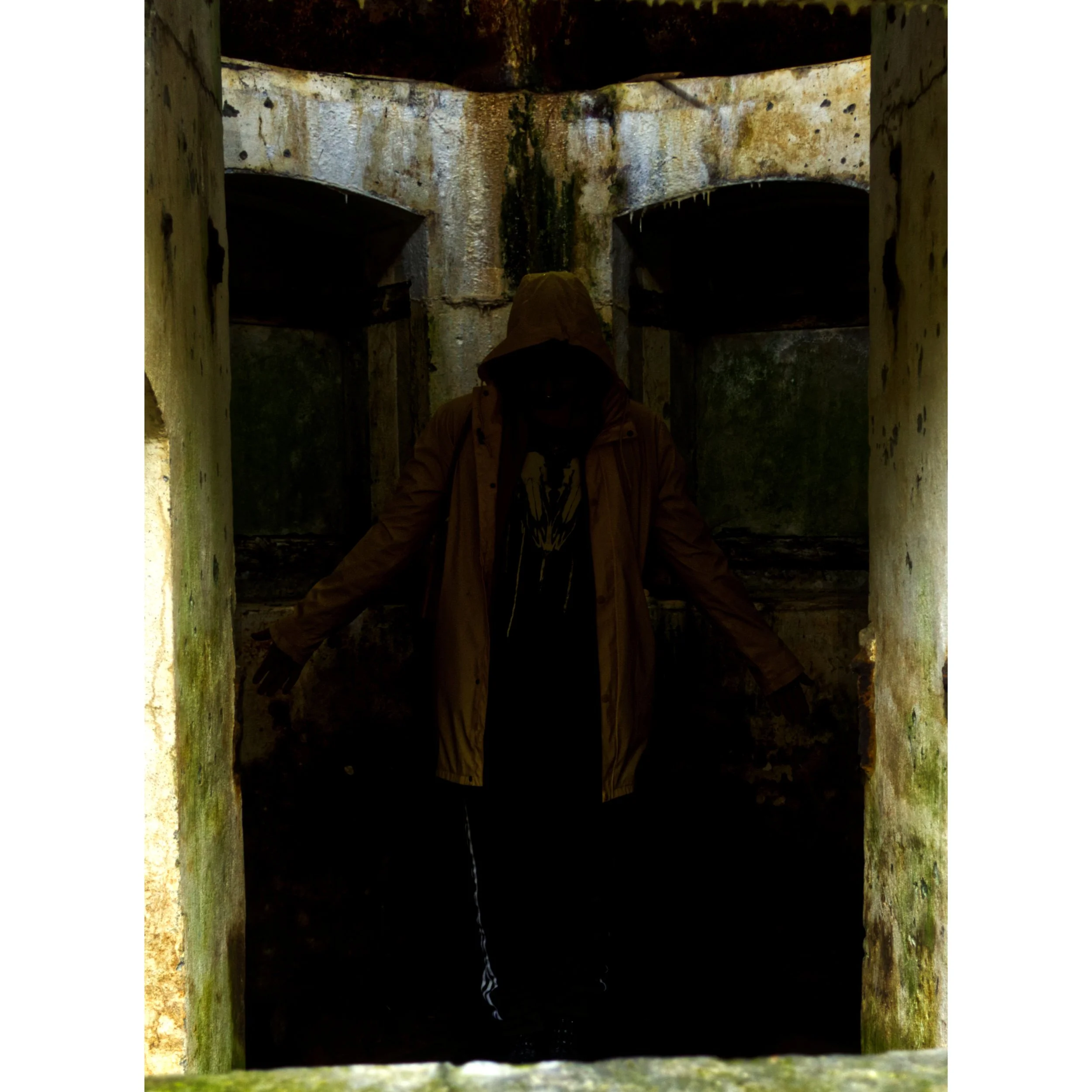 Person wearing a brown hooded jacket standing in front of an old, moss-covered concrete structure with two arches, with their arms outstretched and head slightly bowed.