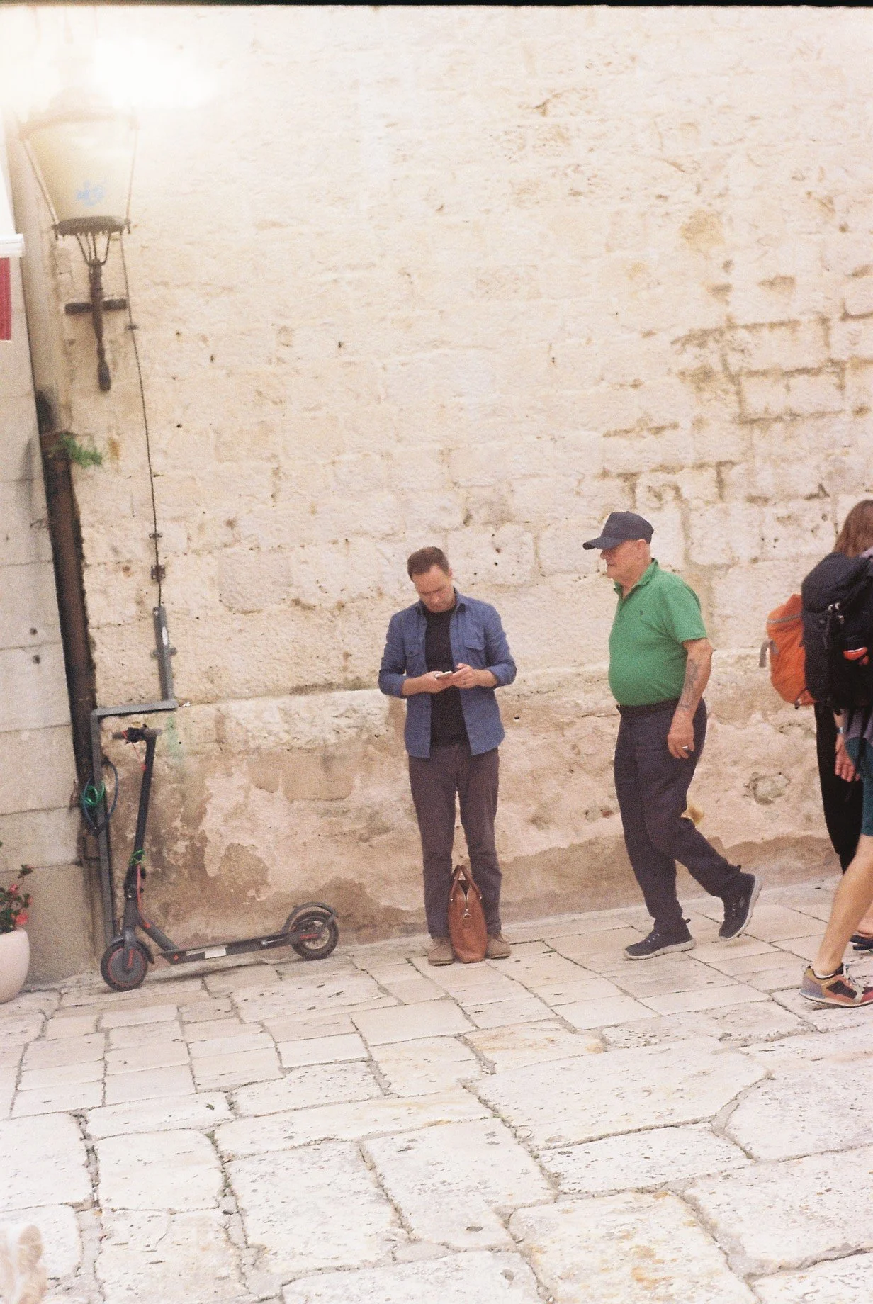 Two men standing on a cobblestone street near a stone wall, one looking at a smartphone, the other walking past, with a scooter leaning against the wall.