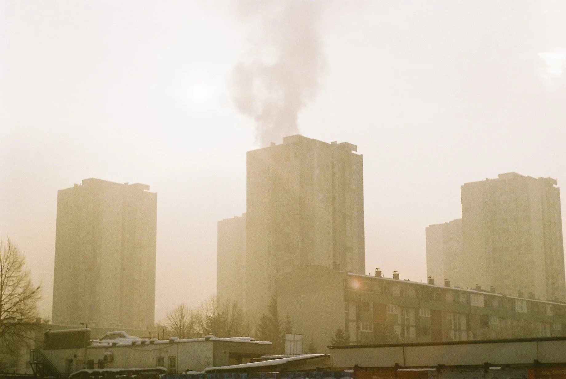 A cityscape with high-rise apartment buildings and thick smoke rising from the top of one building, with a layer of smog or dust creating a hazy atmosphere.