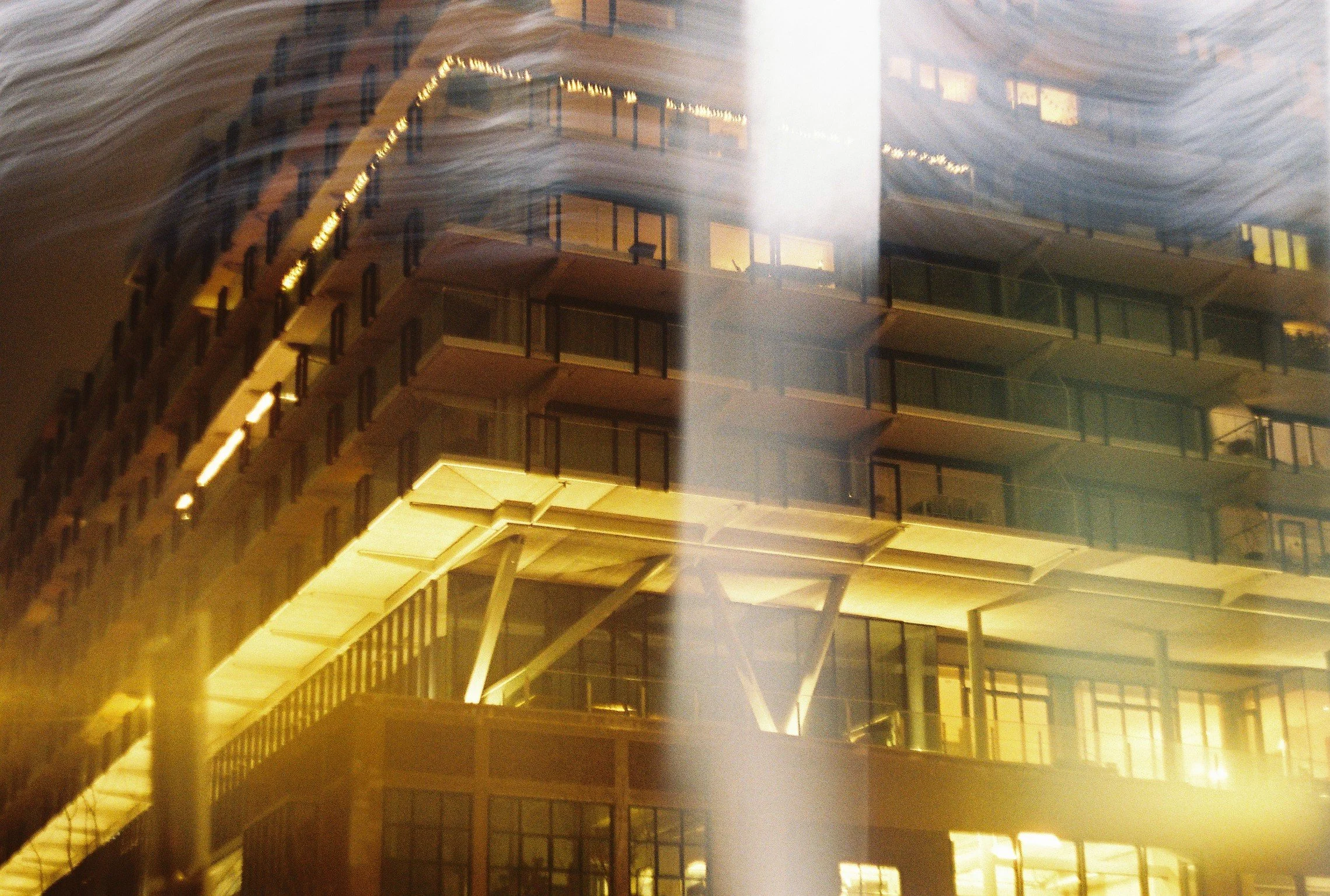 A tall, multi-story building with large glass windows and balconies, illuminated at night with in-built lighting, reflected in water creating a mirror image.