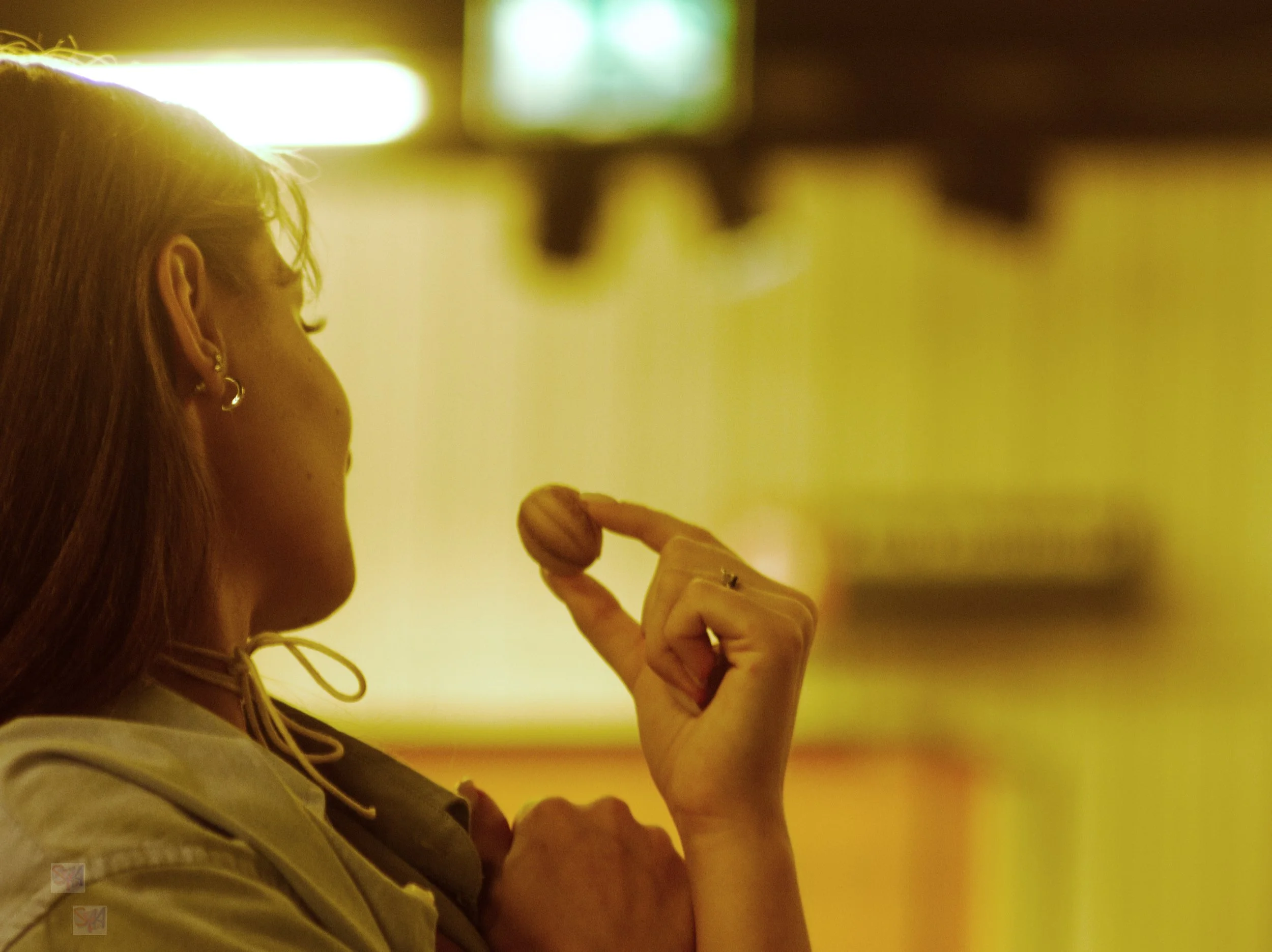 A woman with earrings and a necklace is holding a small object between her thumb and index finger, with her head turned to the left. The background is blurry with bright and colorful lights.