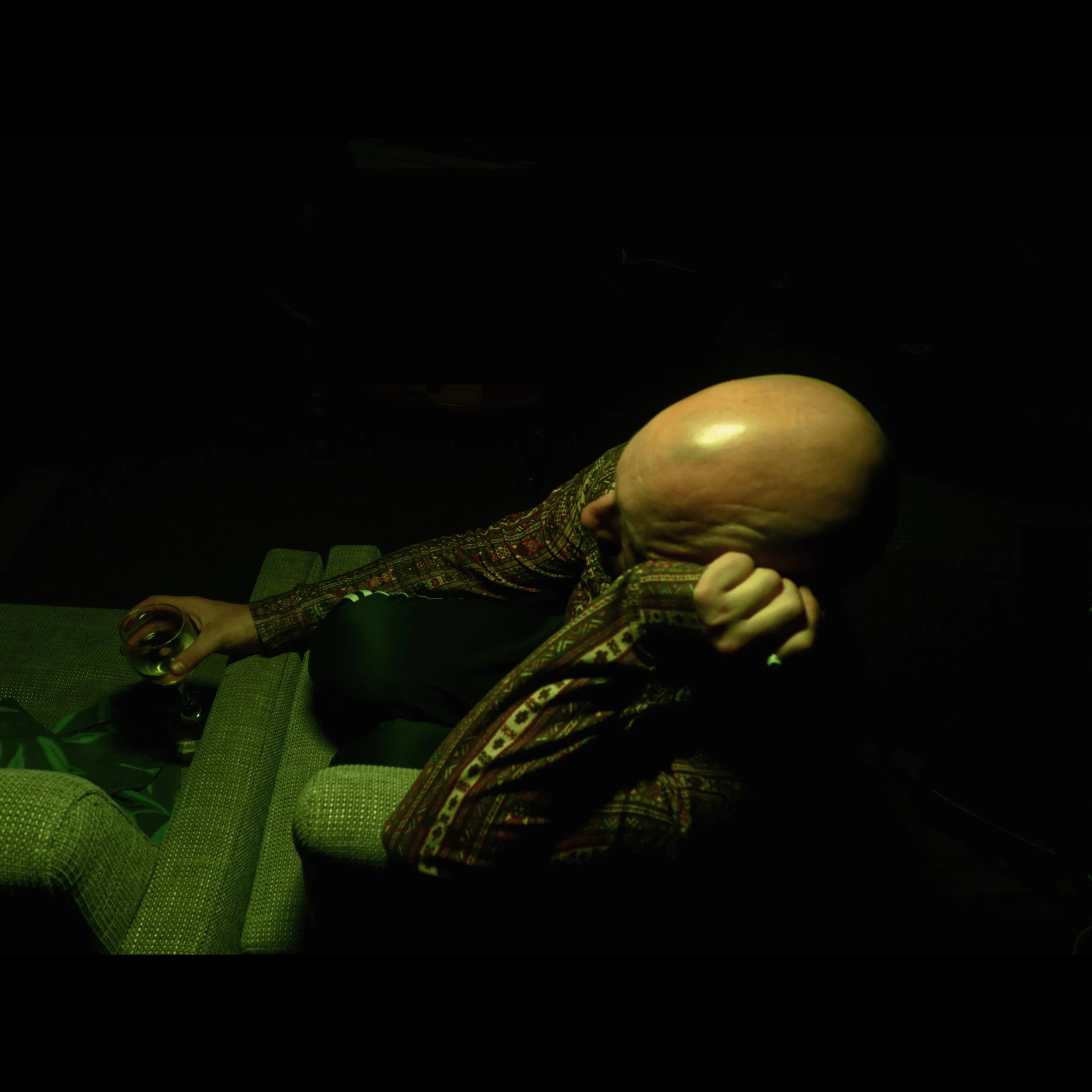 A person with a very large, bald head sitting on a green couch, holding a glass of wine in one hand and resting their face in the other.
