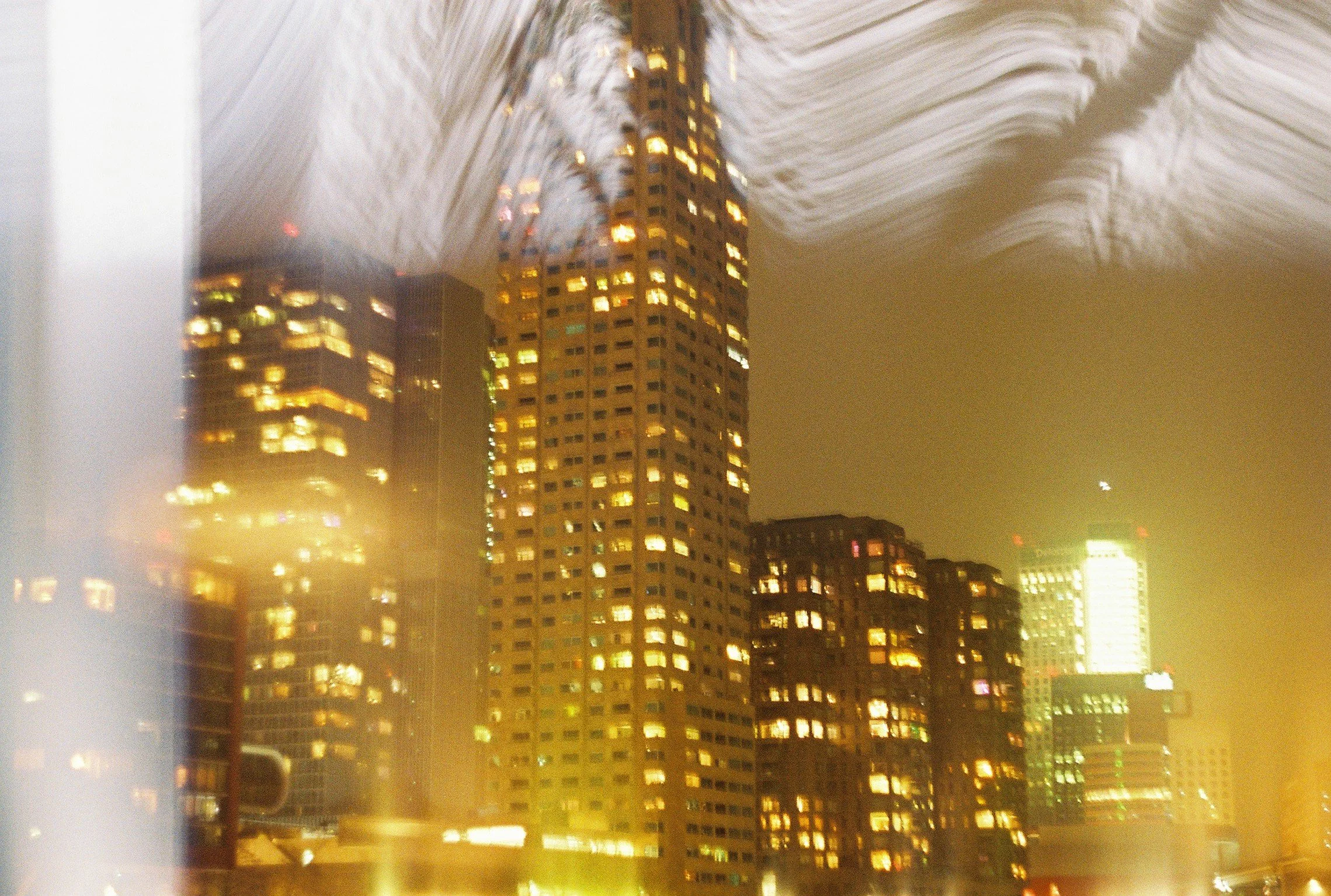 Nighttime cityscape with illuminated skyscrapers, viewed through a rain-covered window with streaks of water and light reflections.