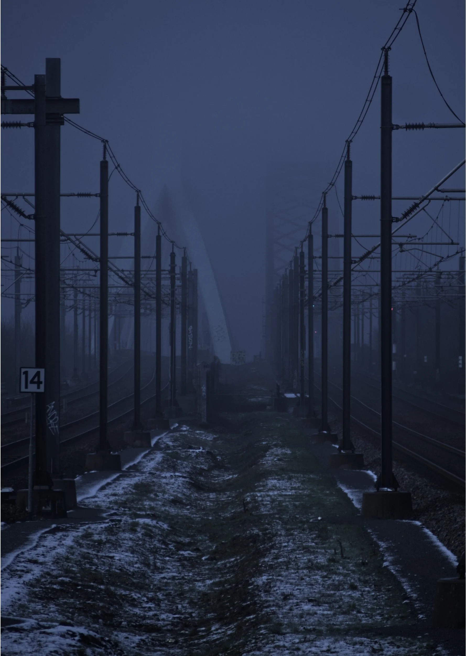 A foggy railway track with electricity poles and lines on either side, with snow on the ground and an industrial cooling tower in the distance.
