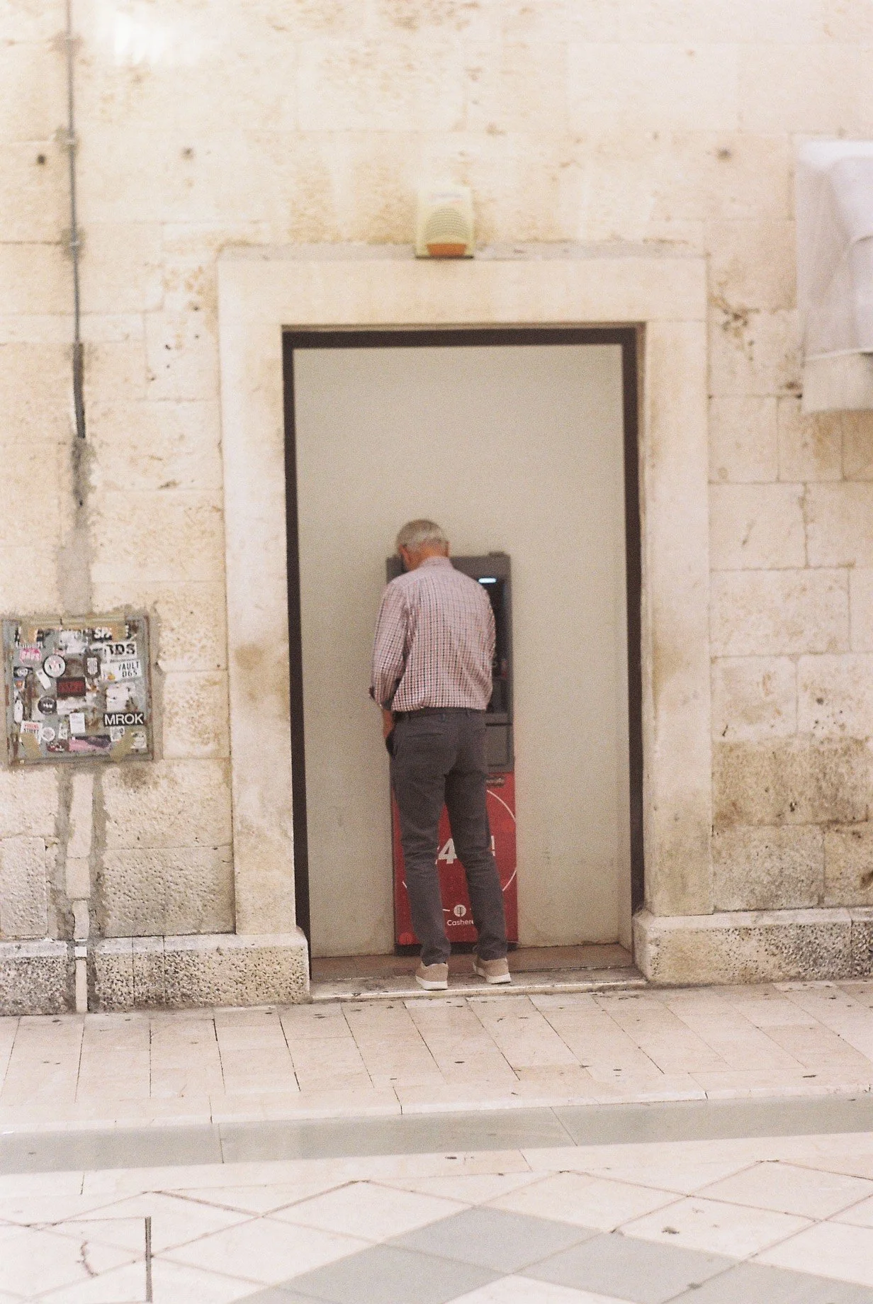 A man with gray hair, wearing glasses, a checkered shirt, and dark pants, stands in front of an ATM inside a building with beige stone walls.