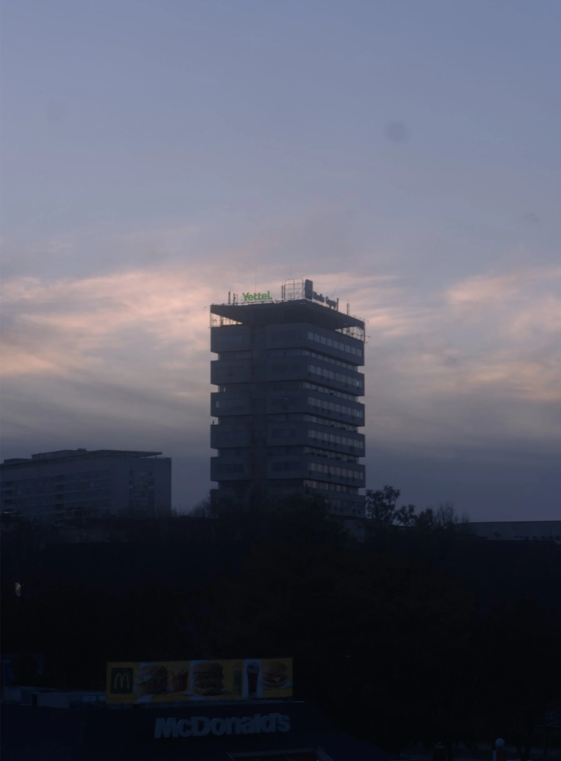 A tall building with a green "Yettel" sign on top, seen against a dusky sky with clouds. A McDonald's sign is visible at the bottom of the image.