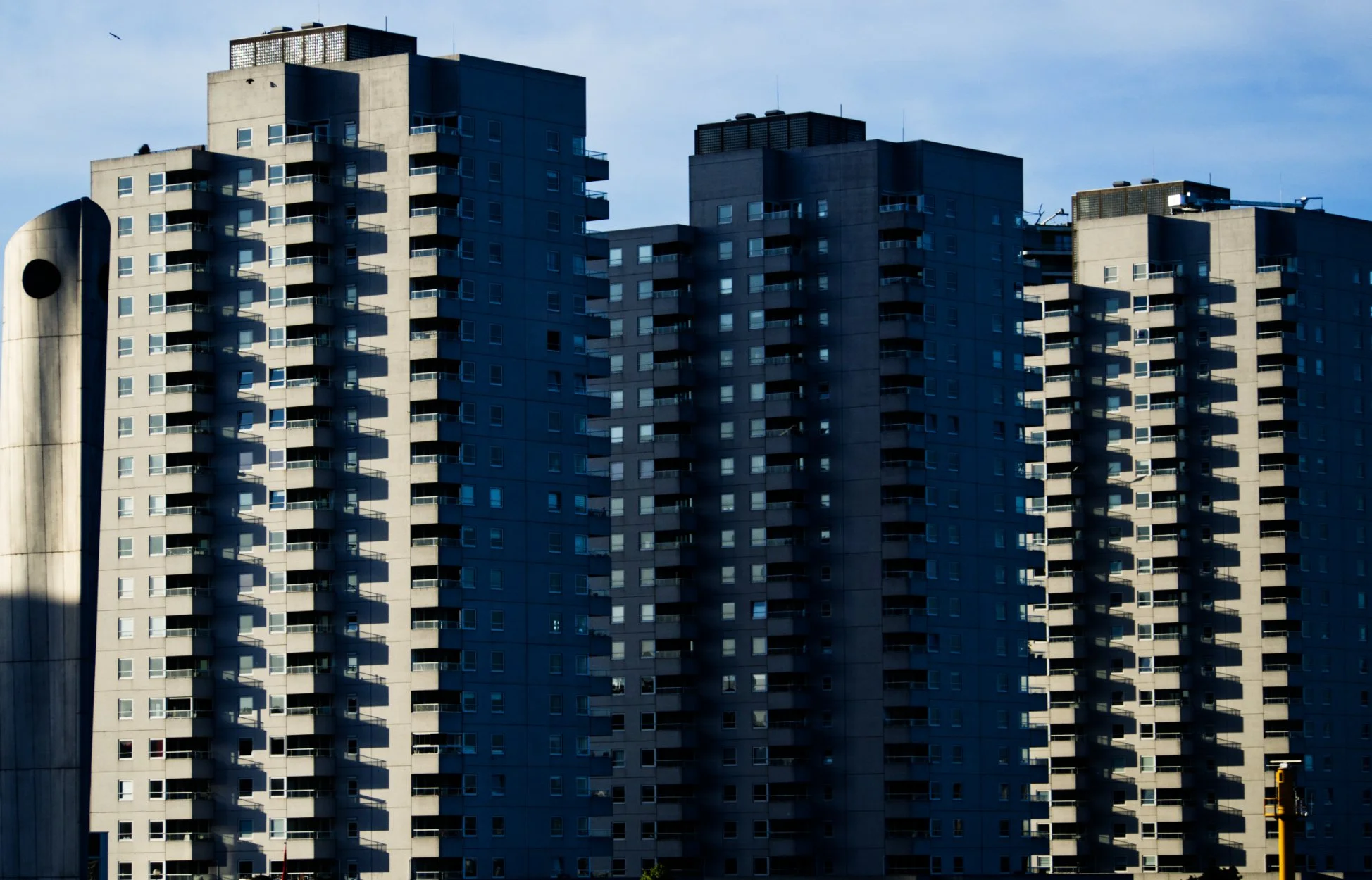 High-rise apartment buildings with numerous windows and balconies, shadows cast by the buildings, and a partly cloudy sky.