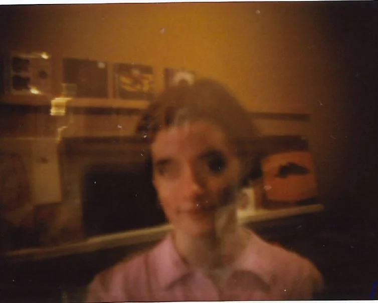 Blurry photo of a woman with short hair, wearing a pink shirt, standing in a room with shelves and various objects in the background.