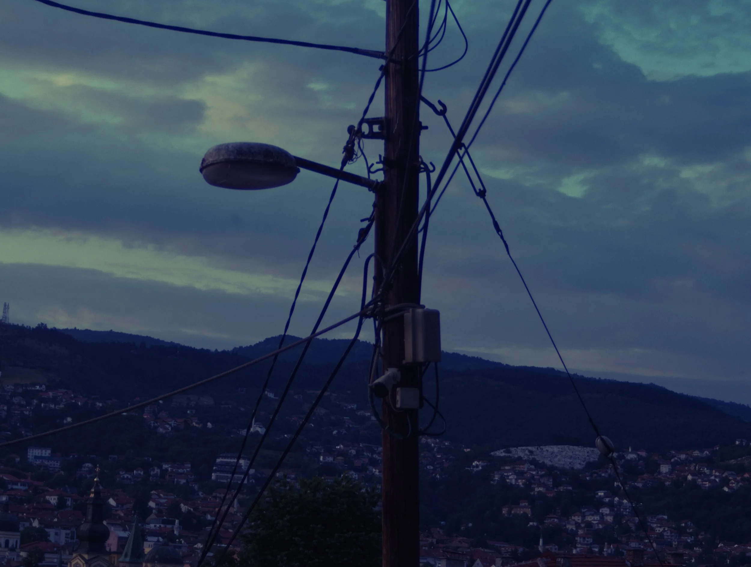 A utility pole with a street light and multiple electrical wires set against a cloudy evening sky above a hillside cityscape.