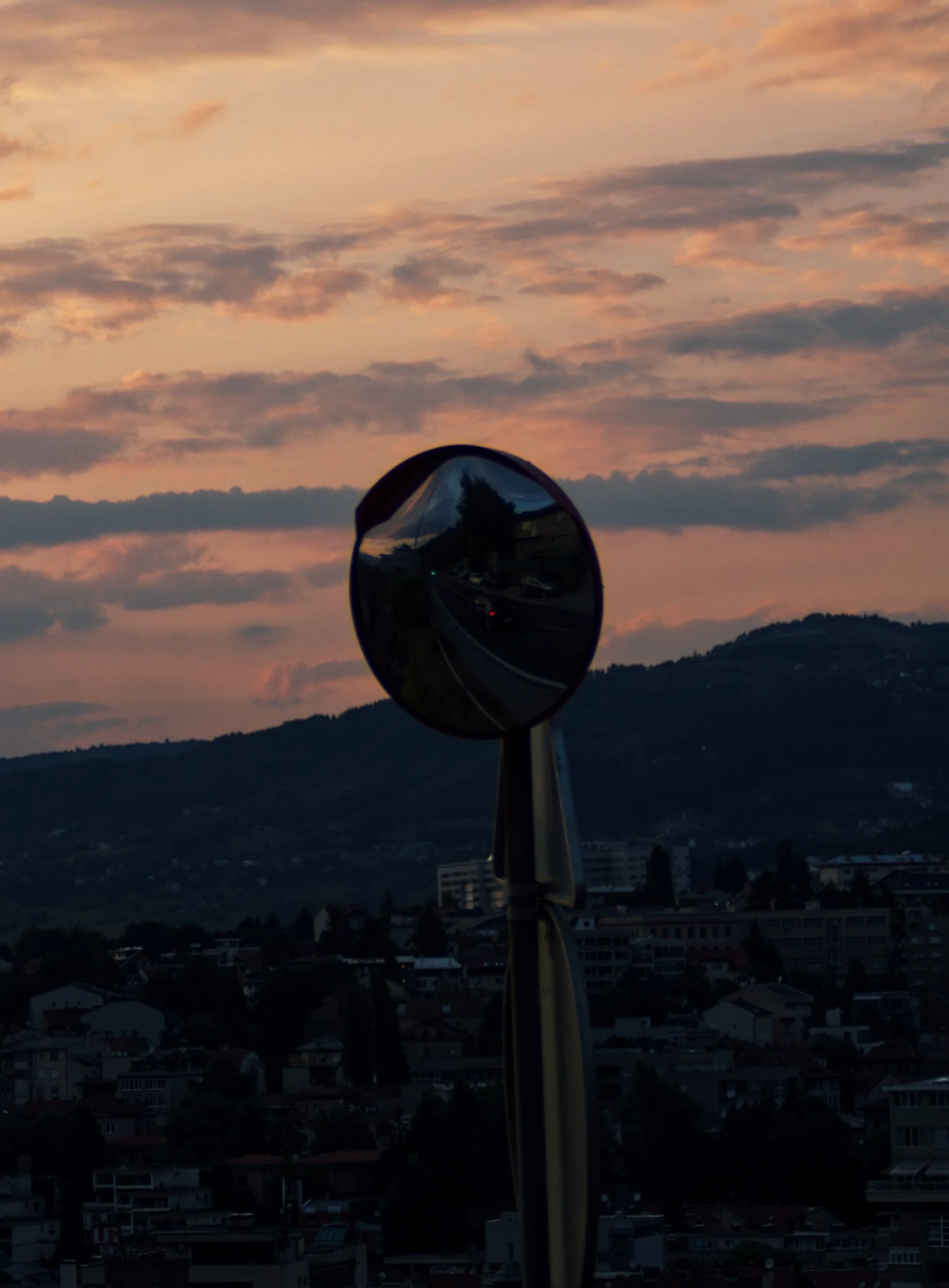 A reflective traffic mirror on a pole captures an urban street scene during sunset, with a mountain and a colorful sky in the background.