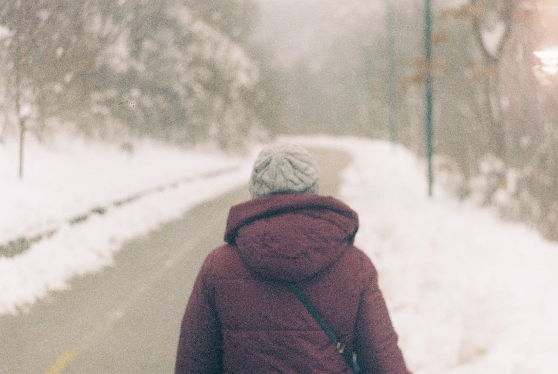 Person walking on a snowy sidewalk at night, wearing a white knit hat and a maroon winter coat.