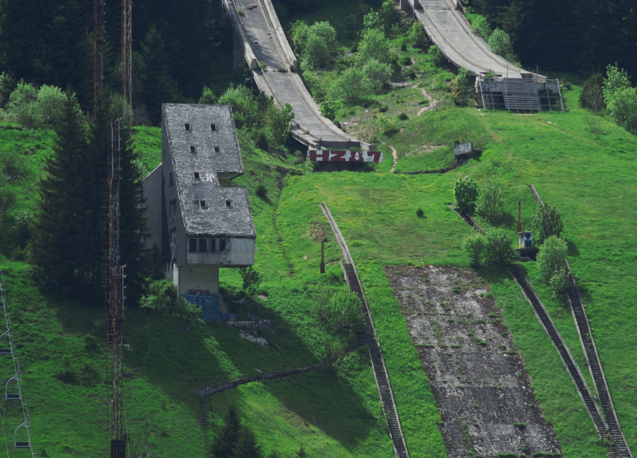 A hillside with chairlifts, a house-like building, and graffiti, surrounded by green trees and grass.