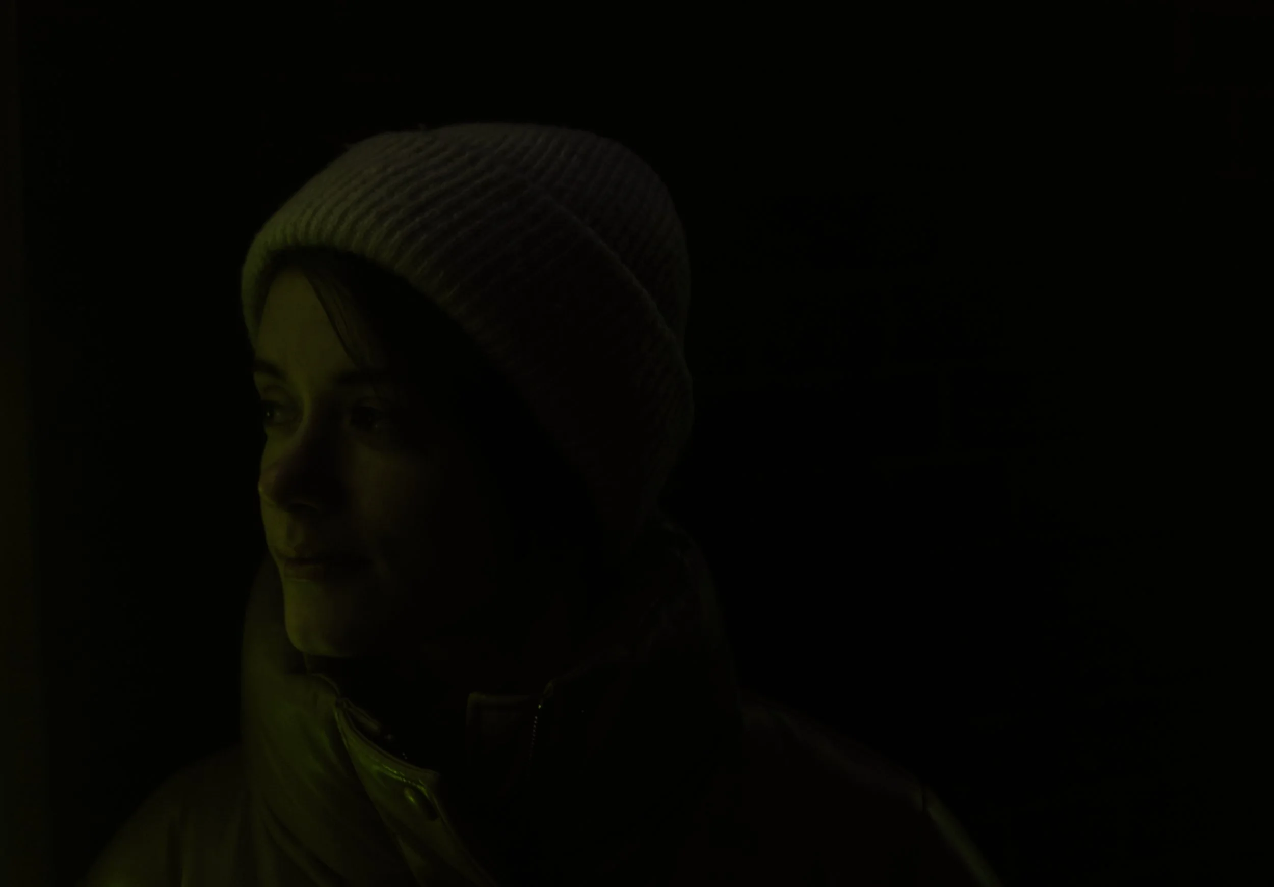 A woman is wearing a knit beanie and a coat, looking sideways in a dimly lit environment.
