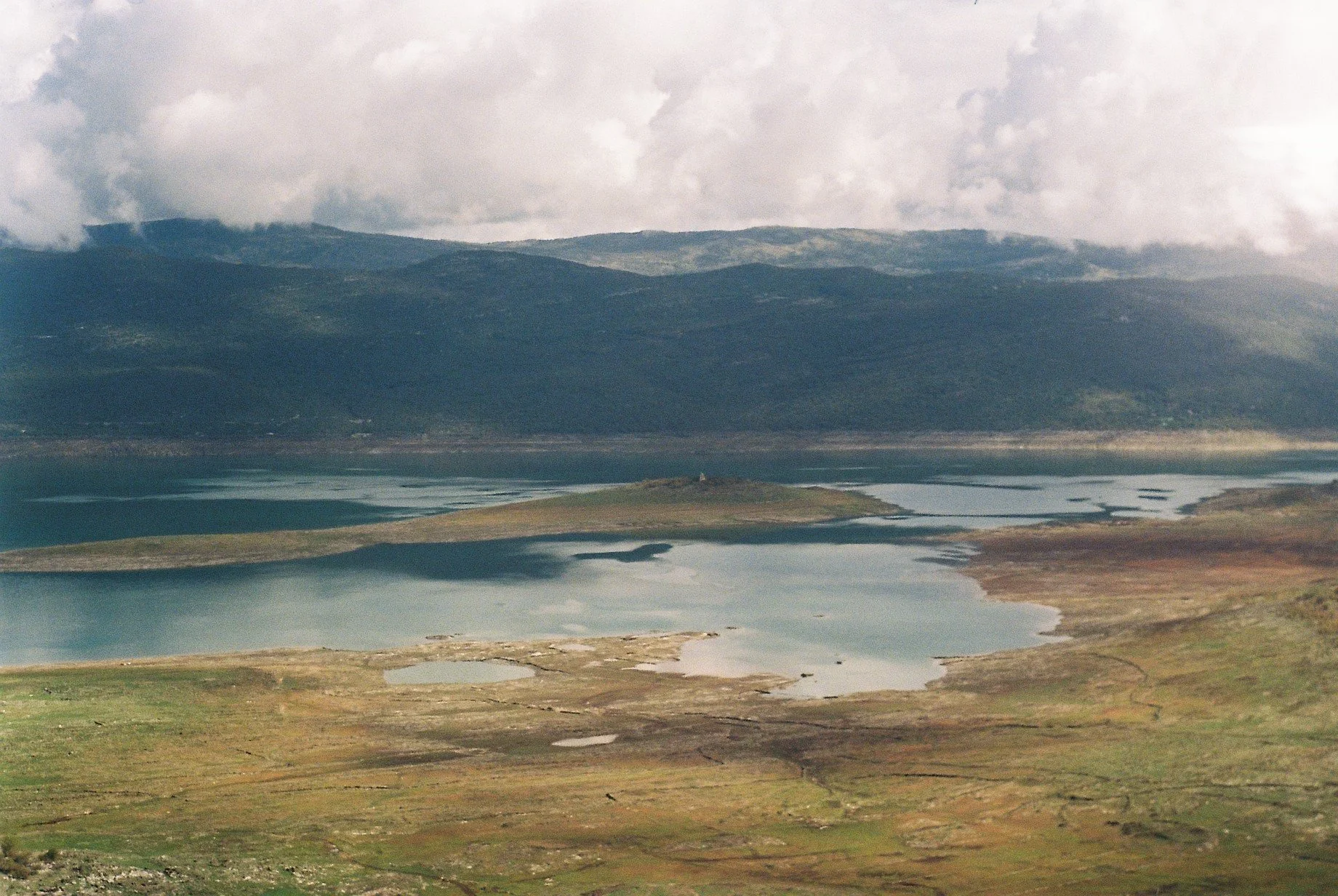 Landscape of a large lake with small islands and surrounding green and brown fields, with mountains and cloudy sky in the background.