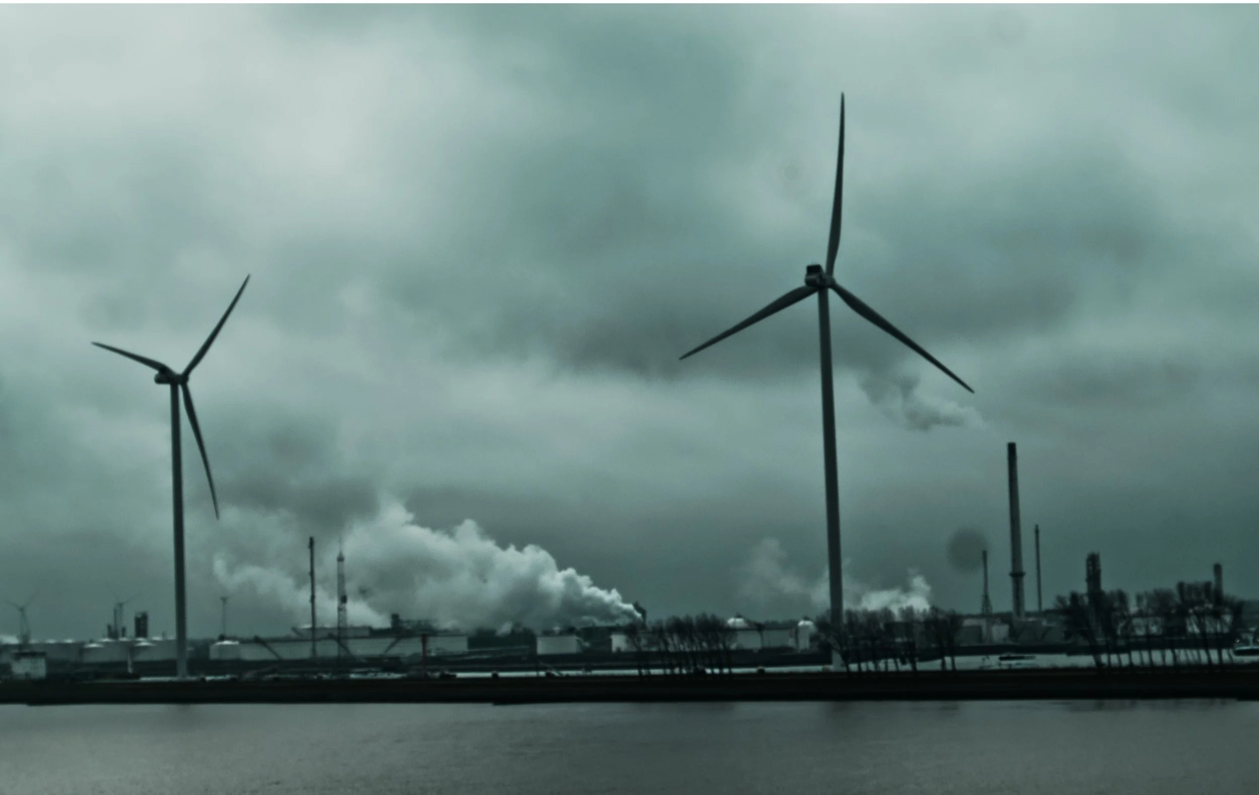 View of wind turbines near an industrial factory emitting smoke and pollution into cloudy sky.