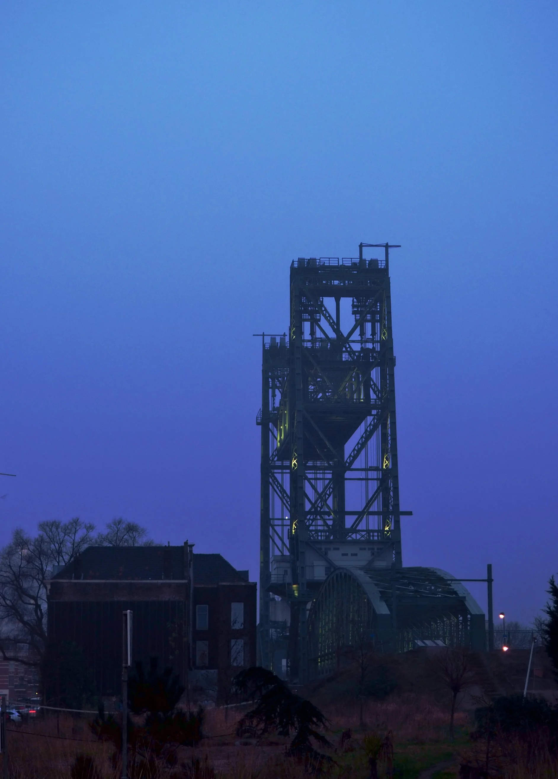 An industrial metallic structure, possibly a mining or oil rig, silhouetted against a deep blue evening sky, with some trees and buildings at the foreground.