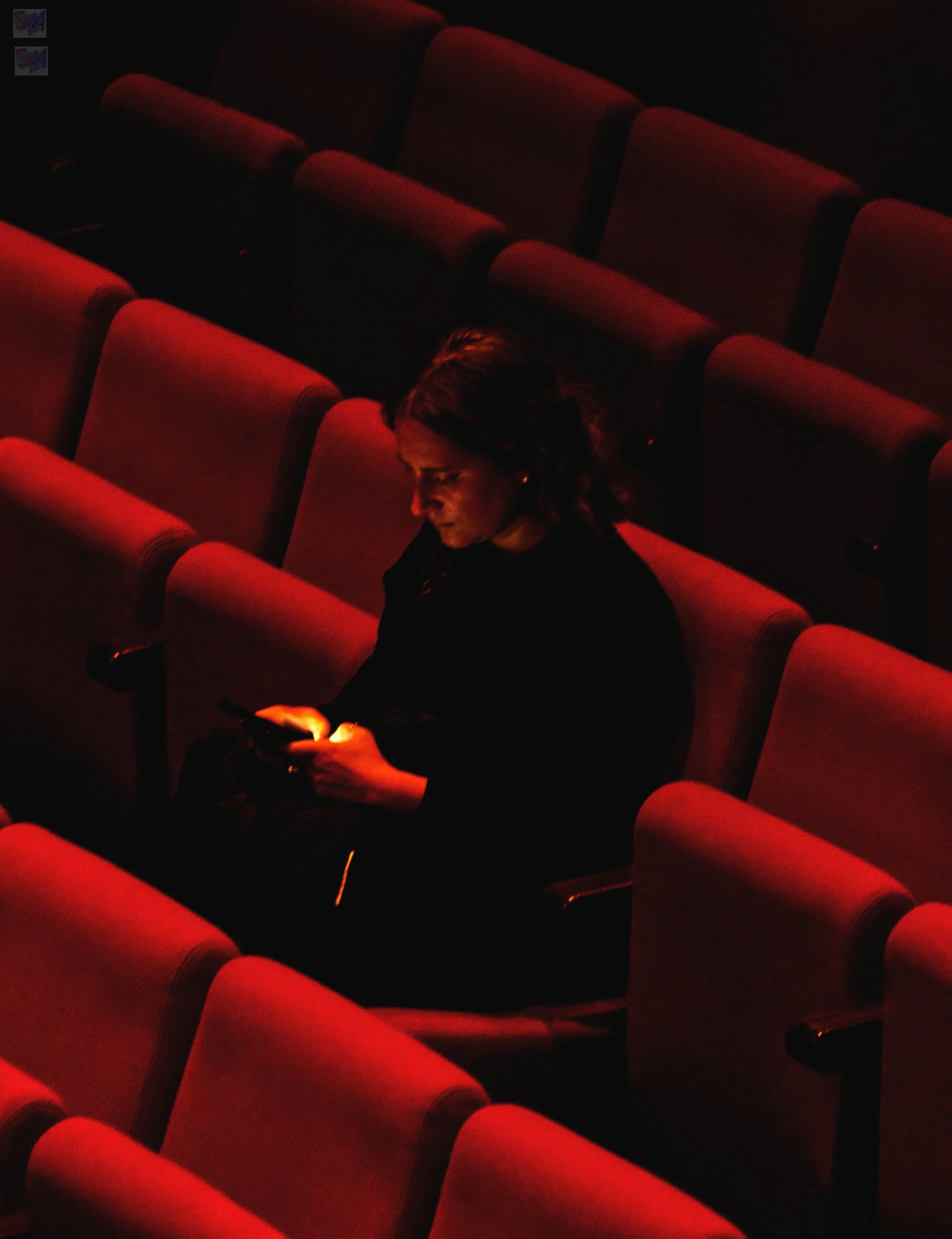 A woman sitting alone in a dimly lit theater with red seats, looking at her phone.