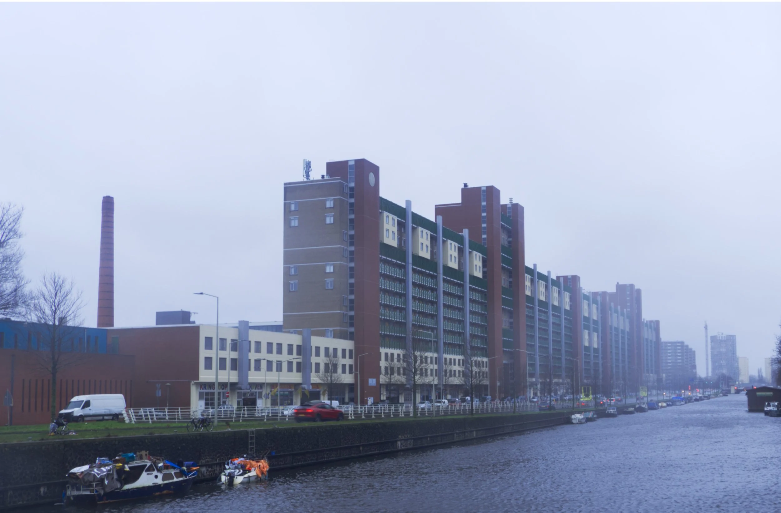 A cityscape with a large modern residential building beside a canal, with boats docked at the edge and a cloudy, foggy sky overhead.