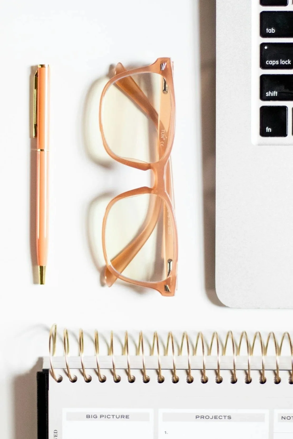 Top view of a workspace with a pink pen, pink glasses, a part of a laptop, and a spiral notebook with labeled sections.