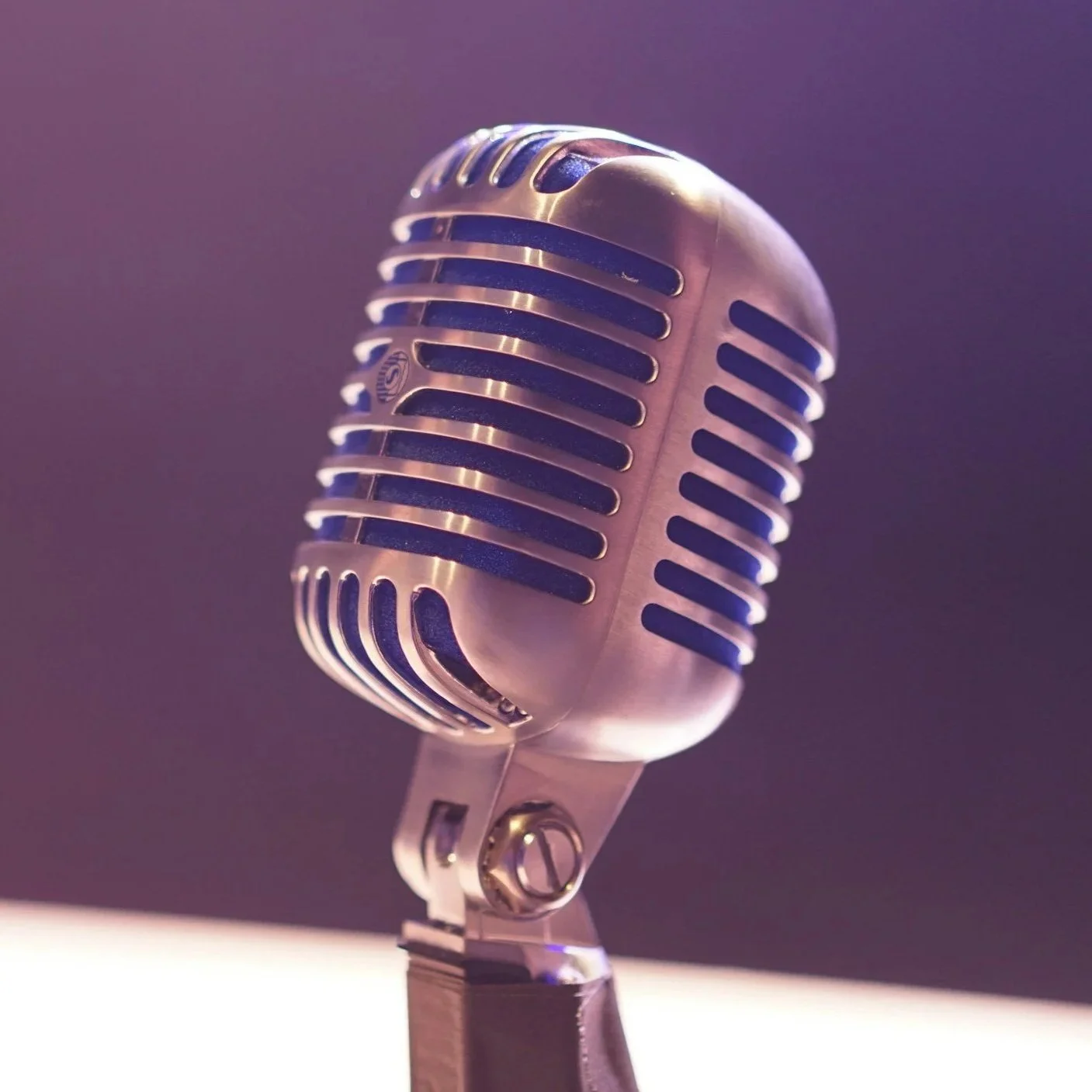 A close-up of a vintage microphone with a metallic grill and stand, with purple lighting in the background.