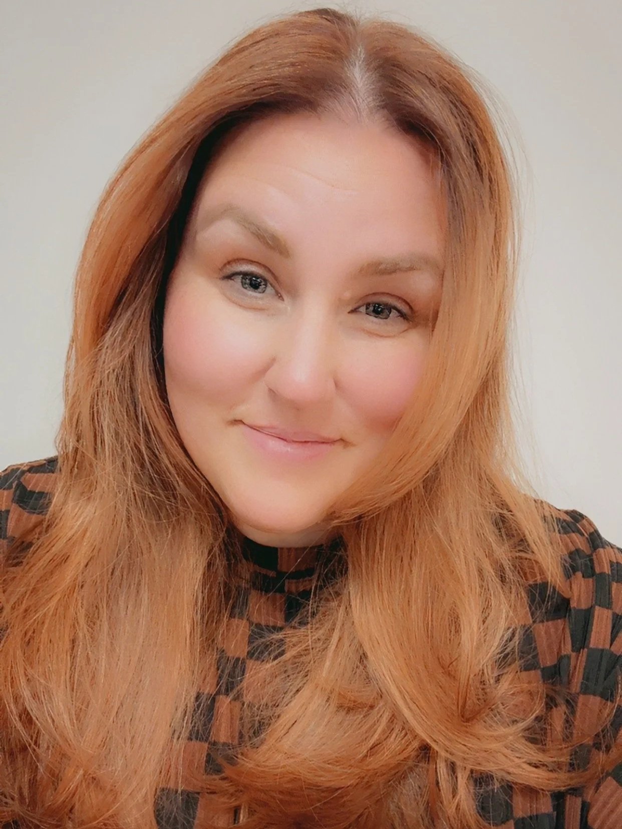 Close-up photo of a smiling woman with long, wavy red hair and fair skin, wearing a checkered shirt with dark and orange tones.