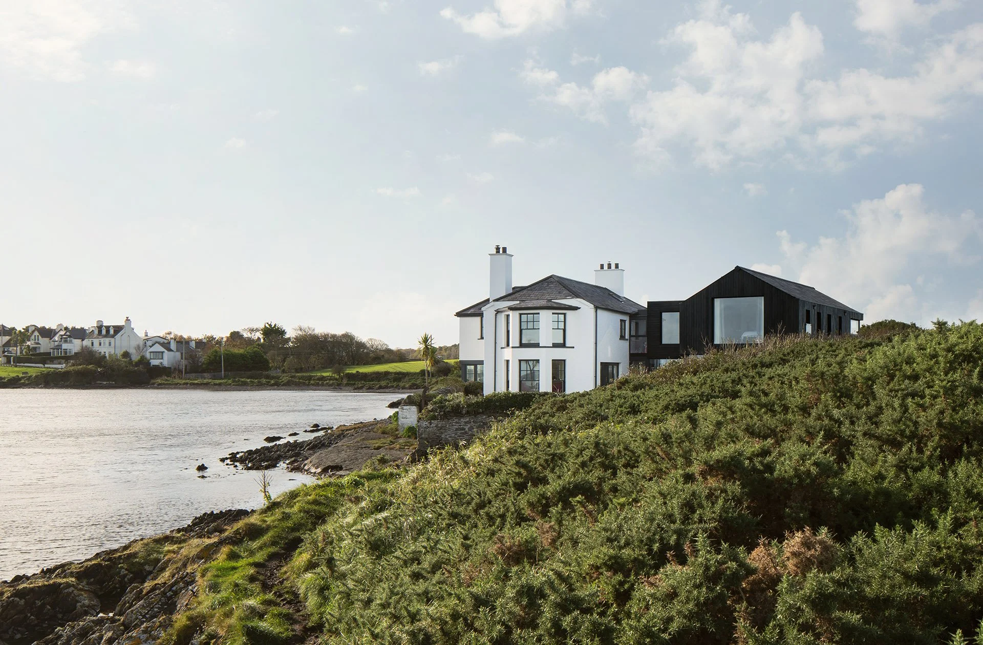 Bradley McClure Architects Ltd — Houses along a waterfront with grassy shoreline, trees, and cloudy sky