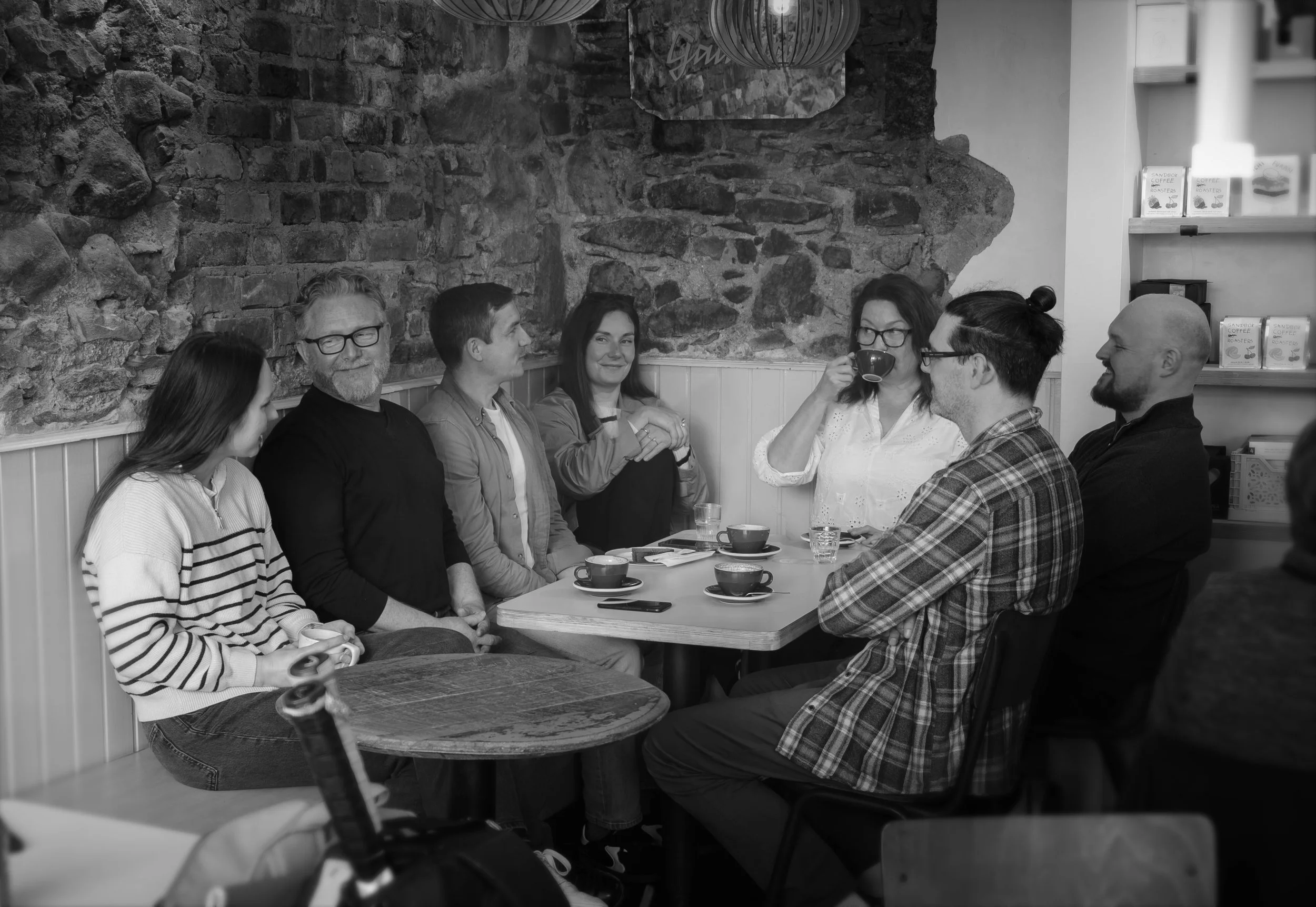 McClure Architects staff team sitting around a table in a cozy cafe, some engaging in conversation or drinking coffee from a mug, against a background of exposed brick wall and shelves with coffee packages.