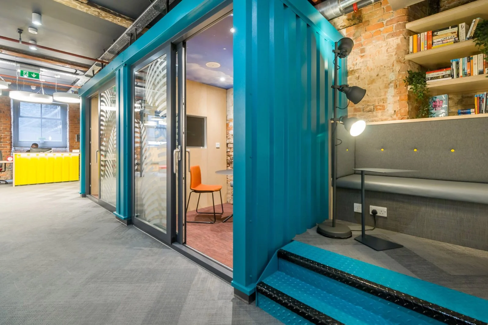 Interior of an office space with colorful decor. There is a glass-walled room with a small orange chair inside. The surrounding area includes a wall with bookshelves, a sectional bench with a table, and modern lighting fixtures. The decor features ex