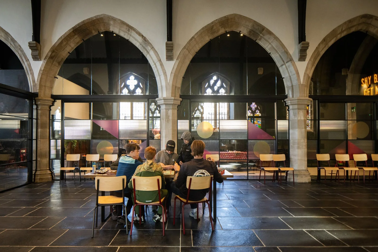 Group of young people sitting at a table inside a building with large arched windows and gothic-style stained glass, engaging in a game or discussion.