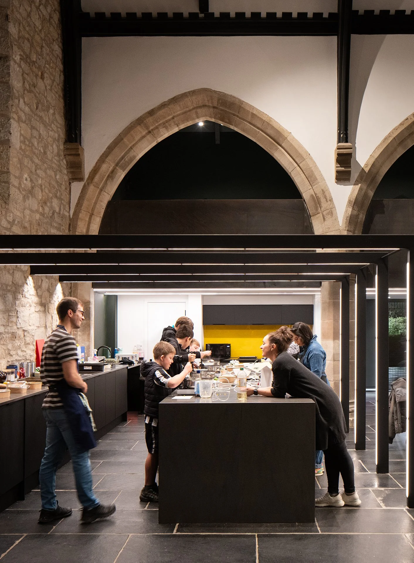 People preparing food and engaging in conversation in a modern, industrial-style kitchen with stone and white walls, high ceilings, and black accents.
