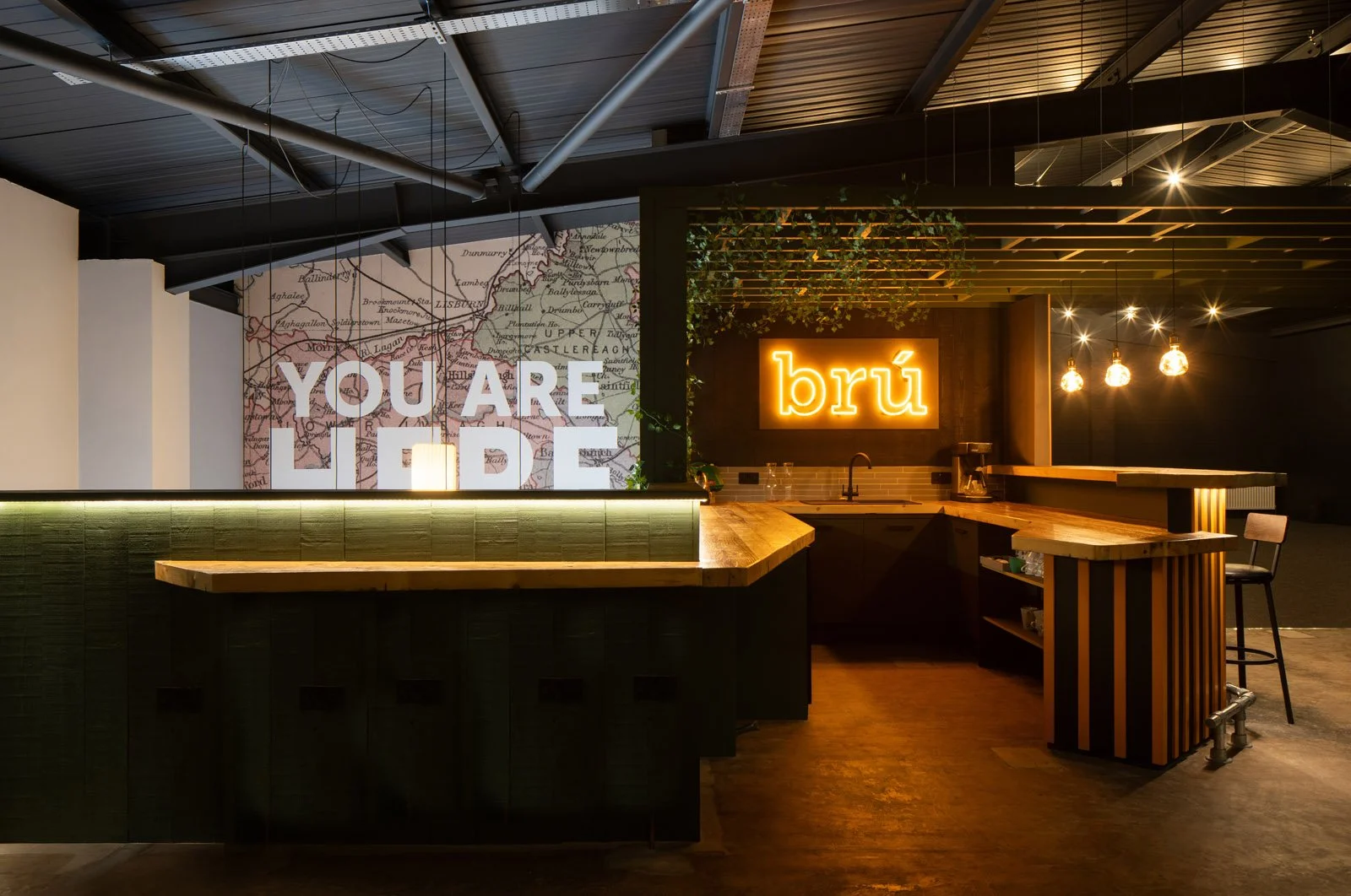 Interior of a modern bar or café with a neon sign that says "brû" and a large wall map with the text "YOU ARE HERE". The space features wooden counters, hanging bulbs, and decorative greenery.