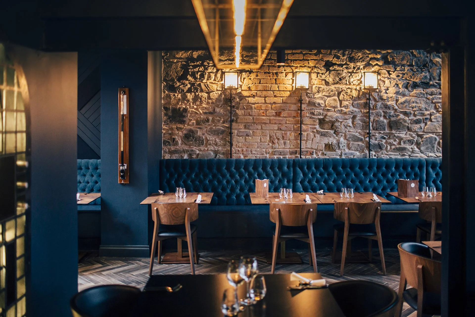 Interior of a restaurant with a brick wall, blue upholstered banquette seating, wooden tables set with wine glasses and menus, and warm lighting fixtures.
