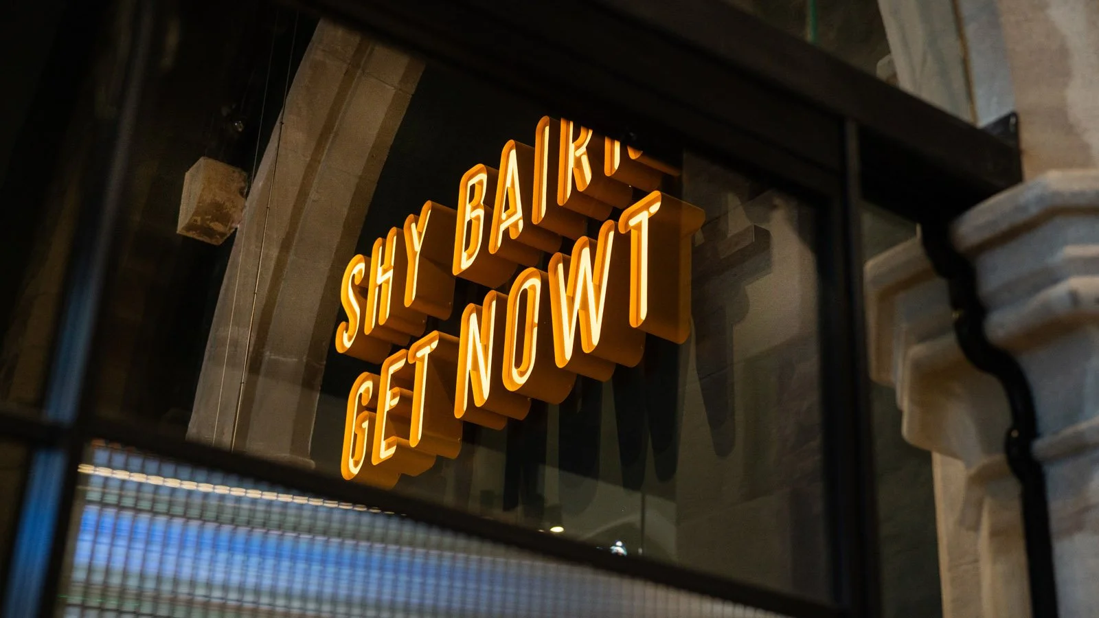 Neon sign that reads 'SHY BAIRNS GET NOWT' attached to a brick wall, inside a building with visible architectural details.