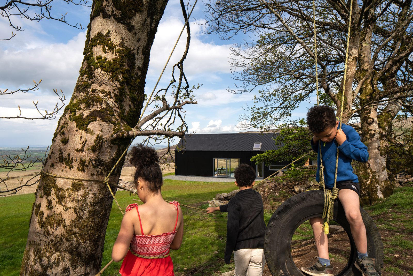 Children playing outdoors near trees, one boy stepping through a large tire, two children holding a rope, and a modern black house in the background.