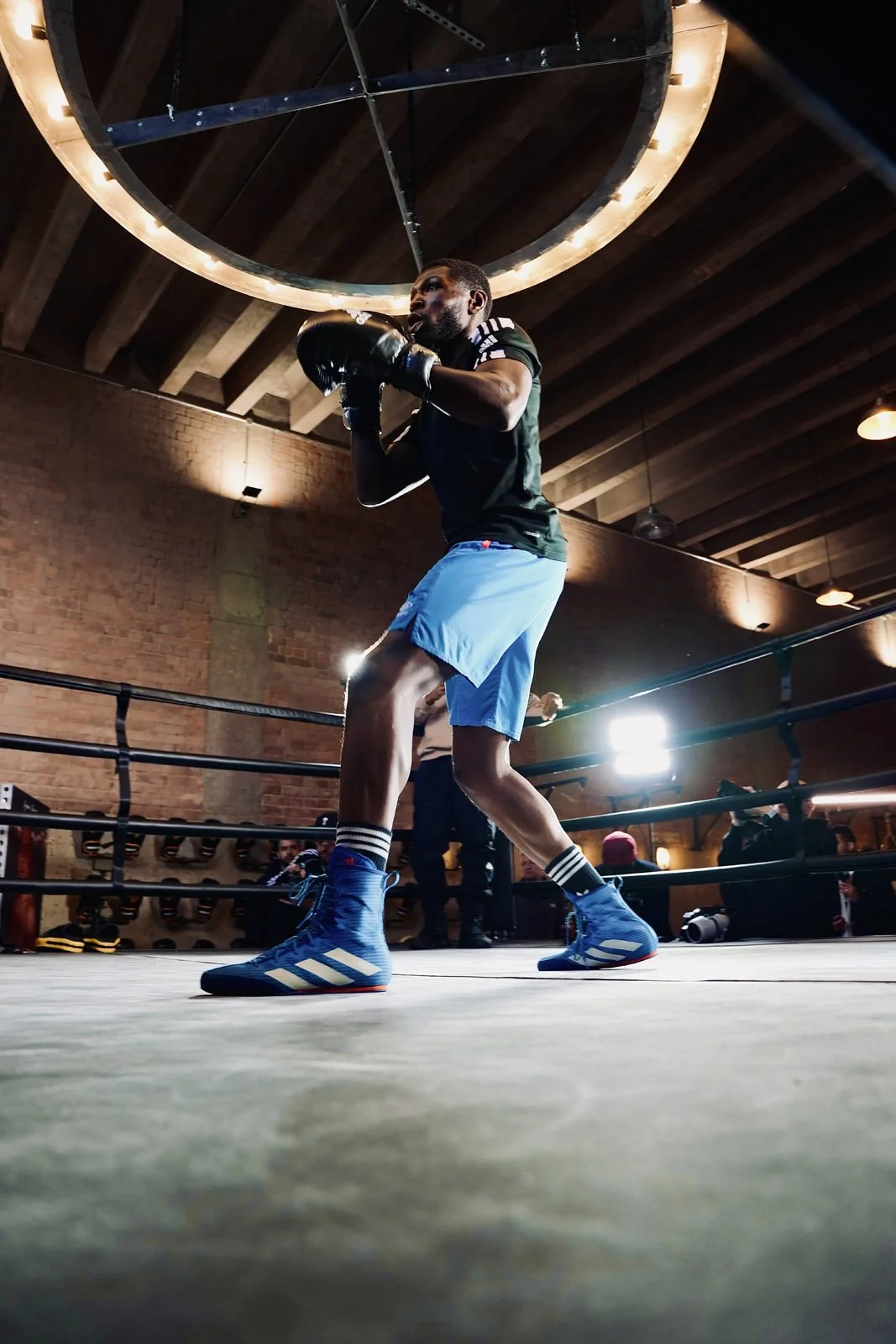 A male boxer in a boxing ring, wearing blue shorts, blue boxing shoes, and black gloves, practicing or sparring under indoor lighting.
