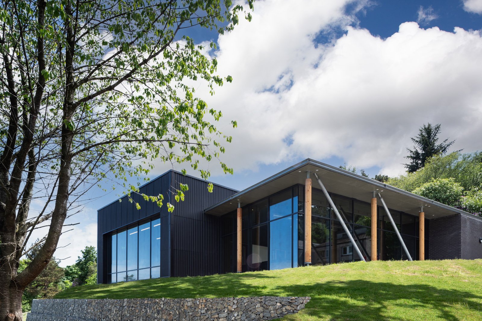 Modern community building with large glass windows, black exterior walls, wooden posts, set on a grassy hill with trees and cloudy sky in the background.