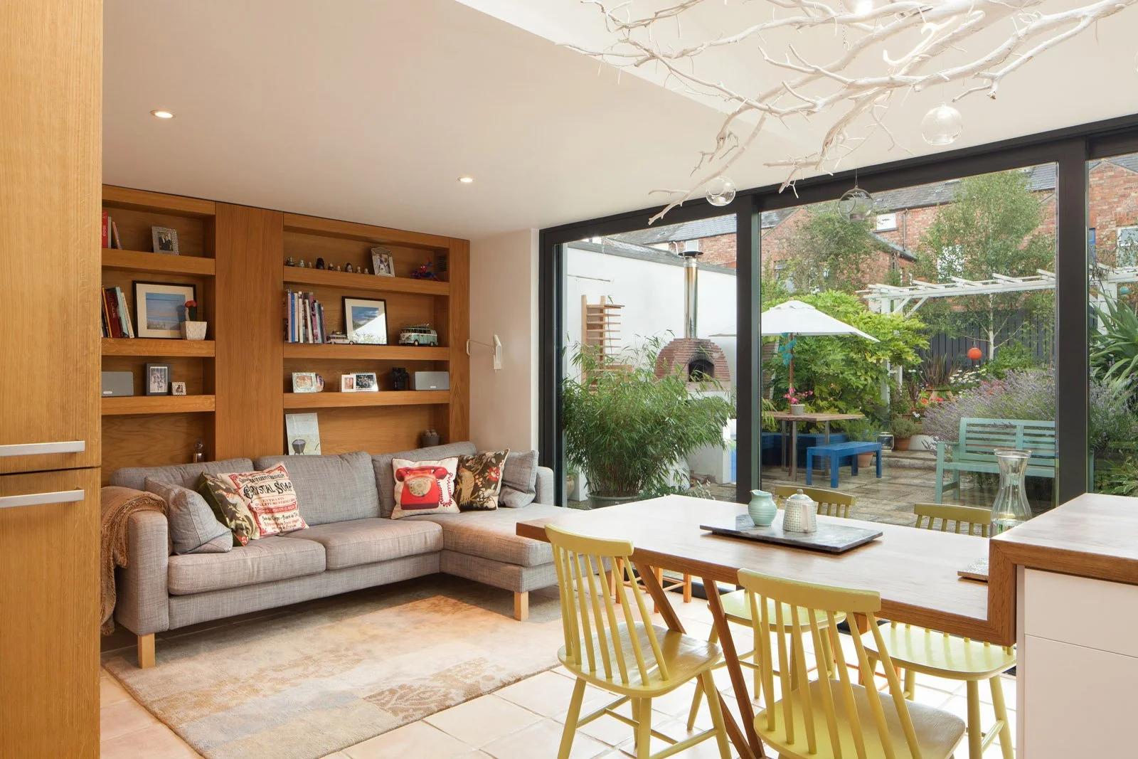 Living room with gray sectional sofa, decorative pillows, wooden bookshelf, large glass sliding doors leading to a backyard patio with outdoor furniture, plants, and a brick oven.