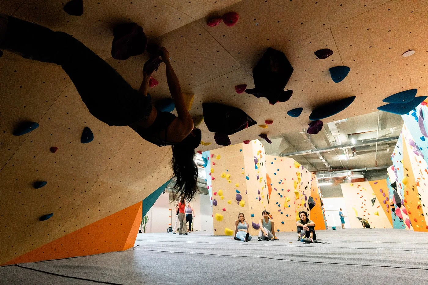 Indoor rock climbing gym with various climbing walls and colorful holds, featuring a person climbing upside down and others sitting or standing nearby.