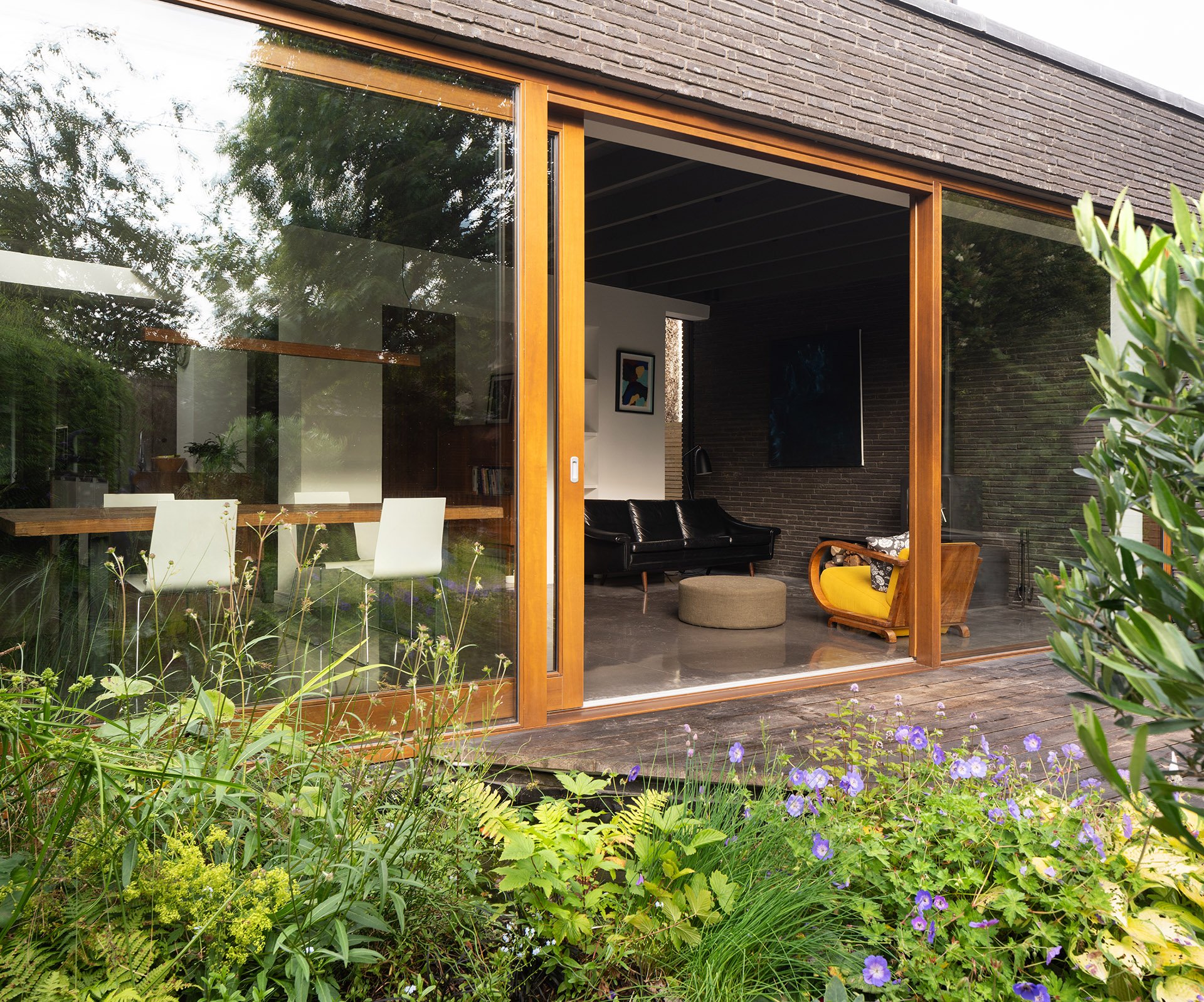 View of a modern house with large glass windows, showing a cozy living room with black leather sofas, artwork on the walls, and a bridge extending over a lush garden with purple flowers and green foliage.