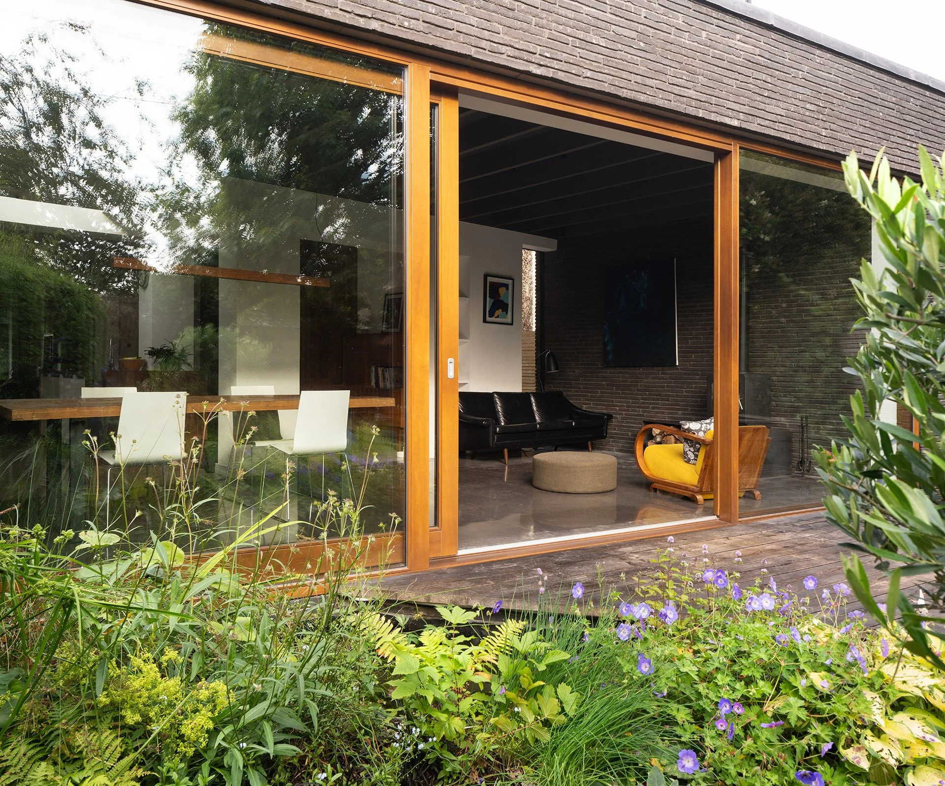 Bradley McClure Architects Ltd — View of a modern house with large glass windows, showing a cozy living room with black leather sofas, artwork on the walls, and a bridge extending ove