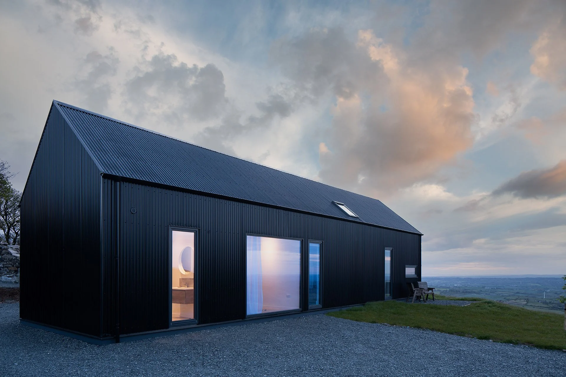 Bradley McClure Architects Ltd — A modern black metal house with large glass windows at dusk, setting on a grassy area with a gravel pathway, with a view of a cloudy sky and distant l