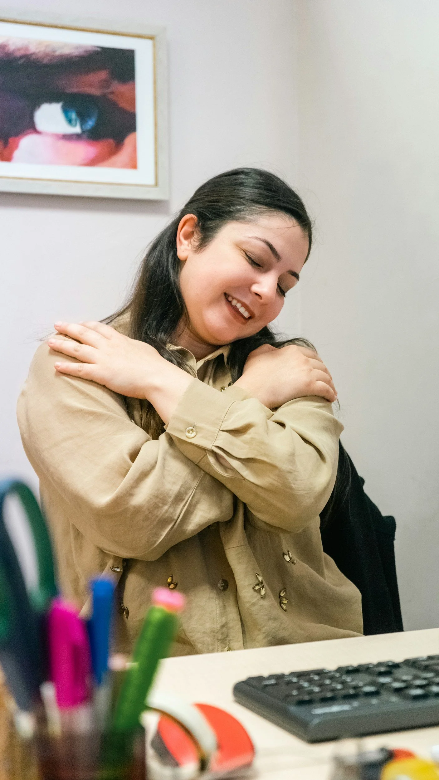 A woman with long dark hair, smiling with closed eyes, hugging herself. She is wearing a beige jacket and sitting at a desk with various office supplies, including colorful pens, a keyboard, and a mouse. A framed picture of an eye is on the wall behind her.